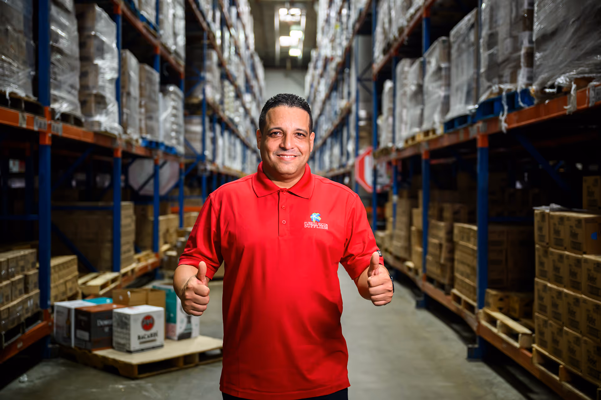 Man in red polo shirt giving two thumbs up standing in a warehouse aisle with shelves stacked with boxes.