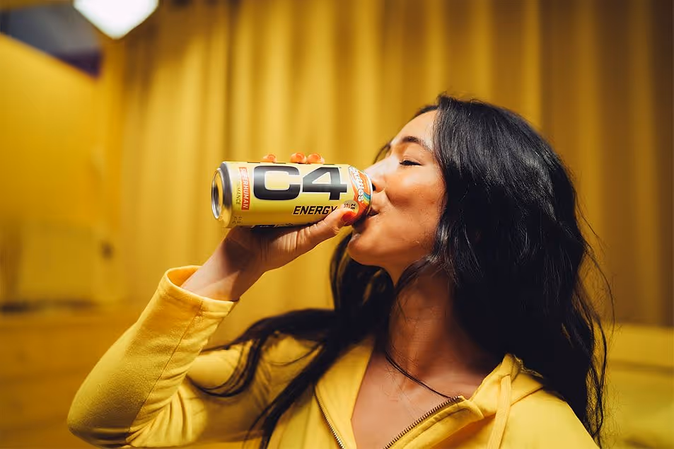 Woman in yellow hoodie drinking a C4 Energy drink against a yellow background.