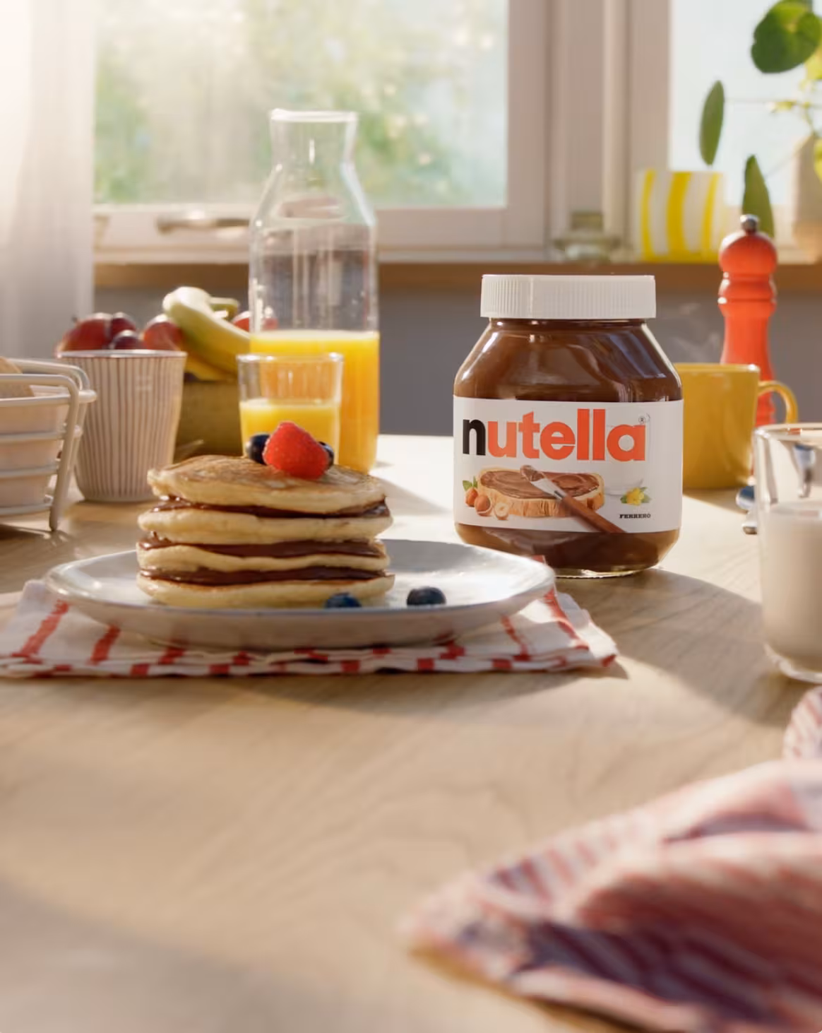 Stack of Nutella-filled pancakes topped with a raspberry and blueberries on a white plate next to a jar of Nutella, orange juice, and breakfast items on a wooden table.