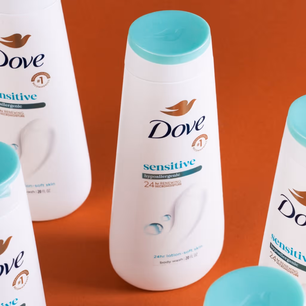 Multiple bottles of Dove Sensitive Hypoallergenic body wash with turquoise caps on an orange background.