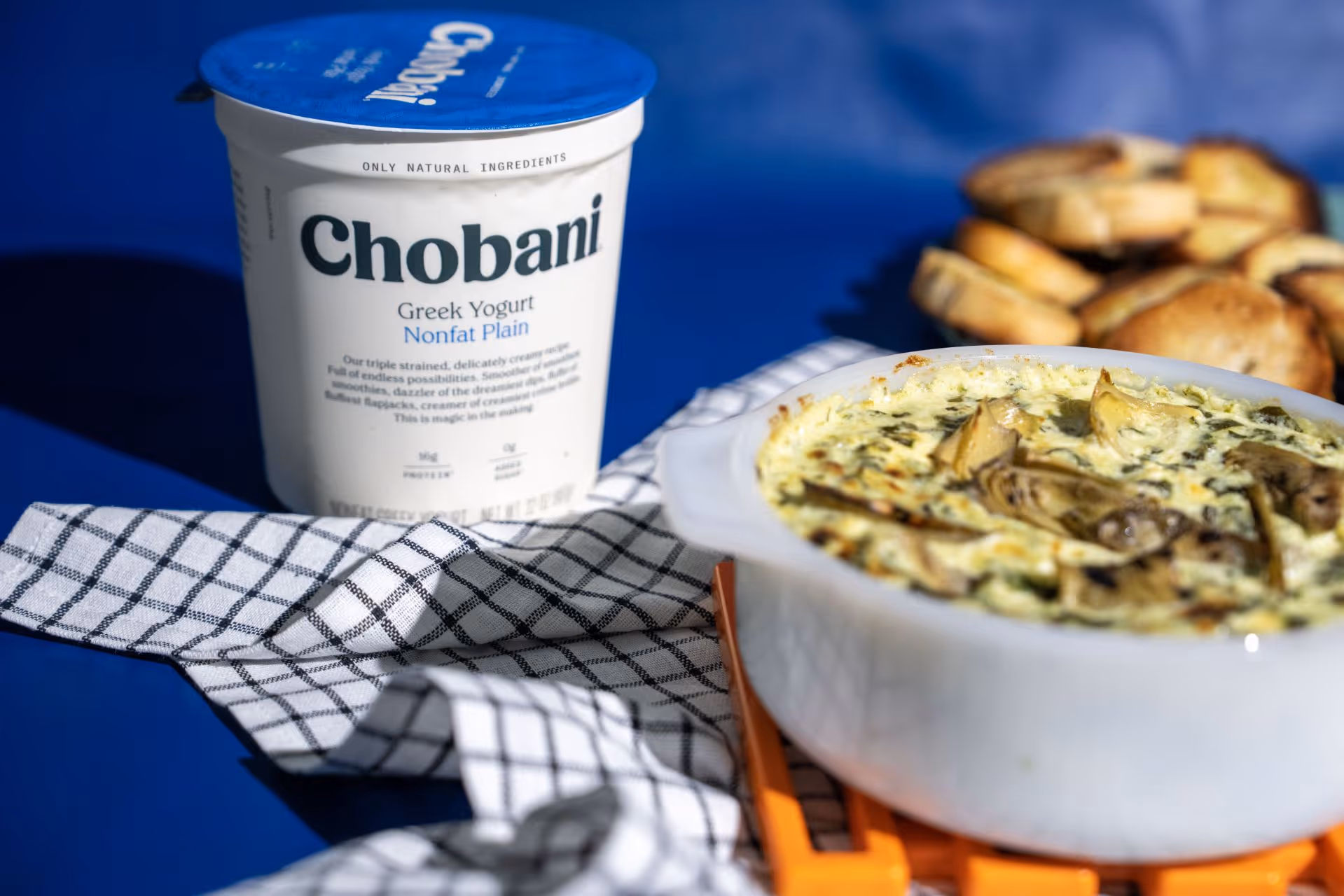 Container of Chobani Nonfat Plain Greek Yogurt next to a white dish of baked spinach and artichoke dip with toasted bread slices in the background.