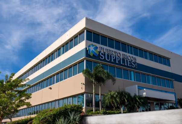 Modern multi-story office building with blue-tinted windows and a sign reading Puerto Rico Supplies Co, surrounded by palm trees and greenery under a partly cloudy sky.