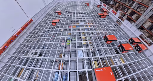 Top view of a modern automated warehouse grid system with robotic shuttles moving red storage bins.