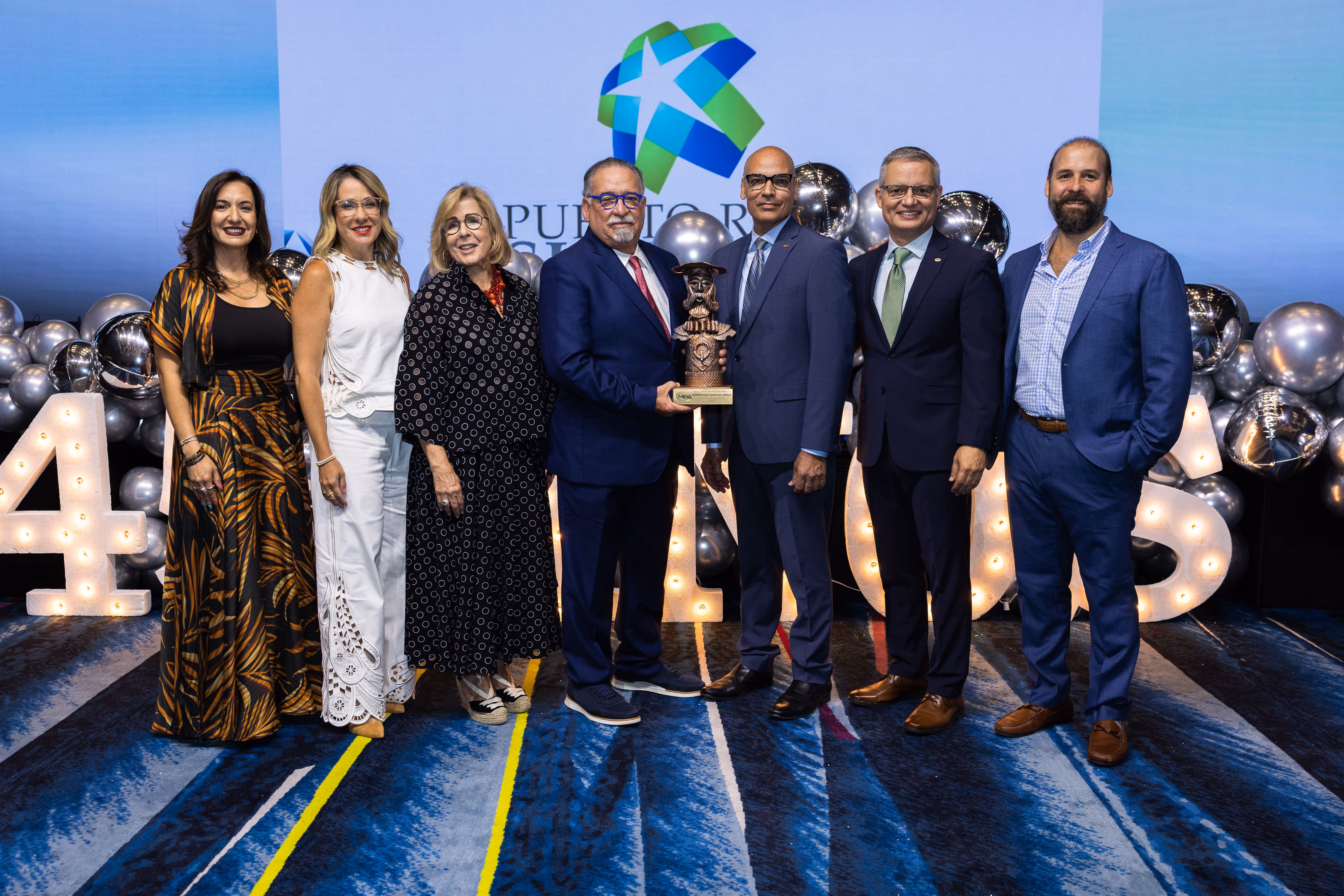 Group of seven people dressed formally standing on a blue carpet in front of illuminated numbers and silver balloons, with one man holding an award trophy.