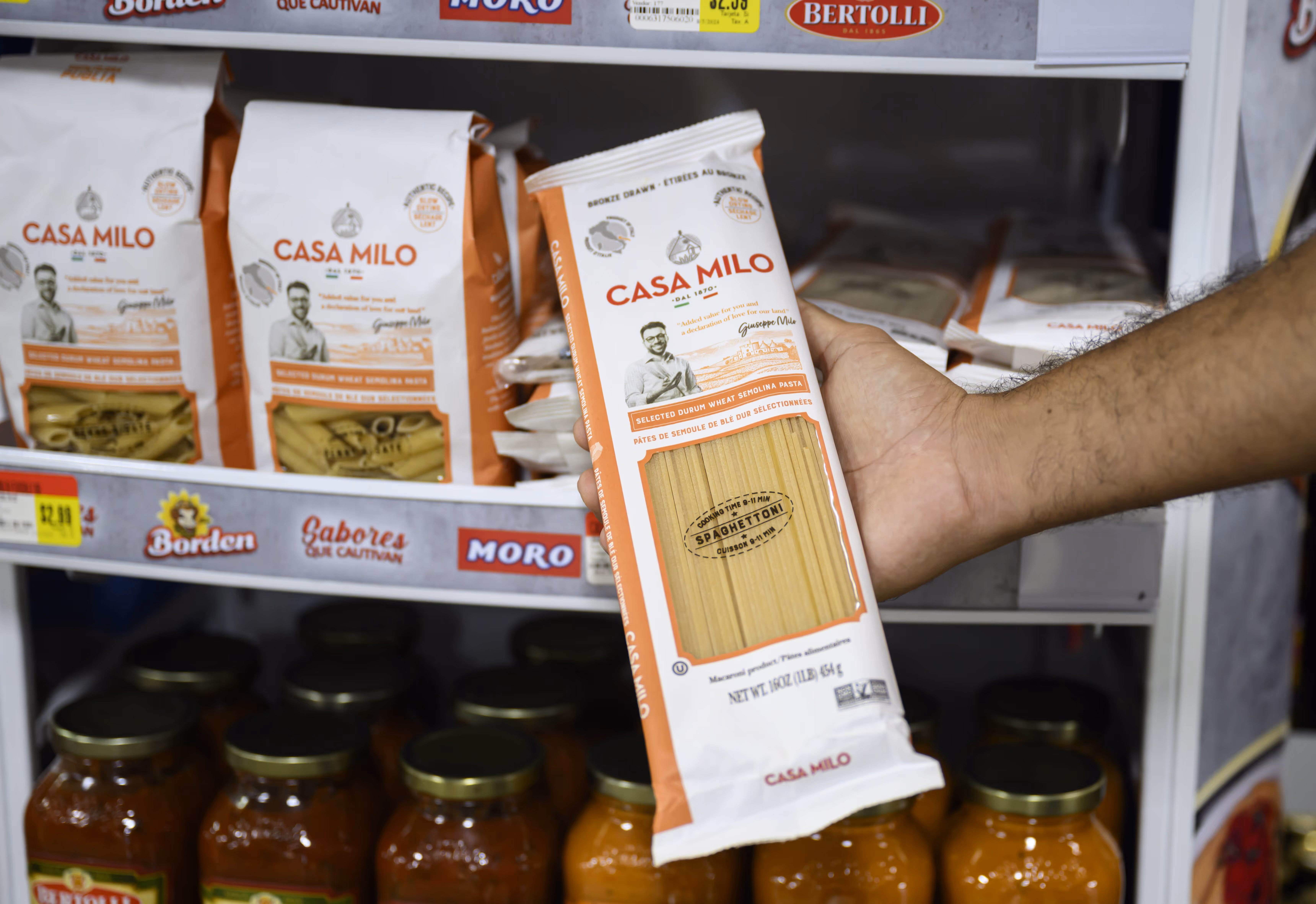 Hand holding a package of Casa Milo spaghetti pasta in a grocery store aisle with more packages and jars on shelves.