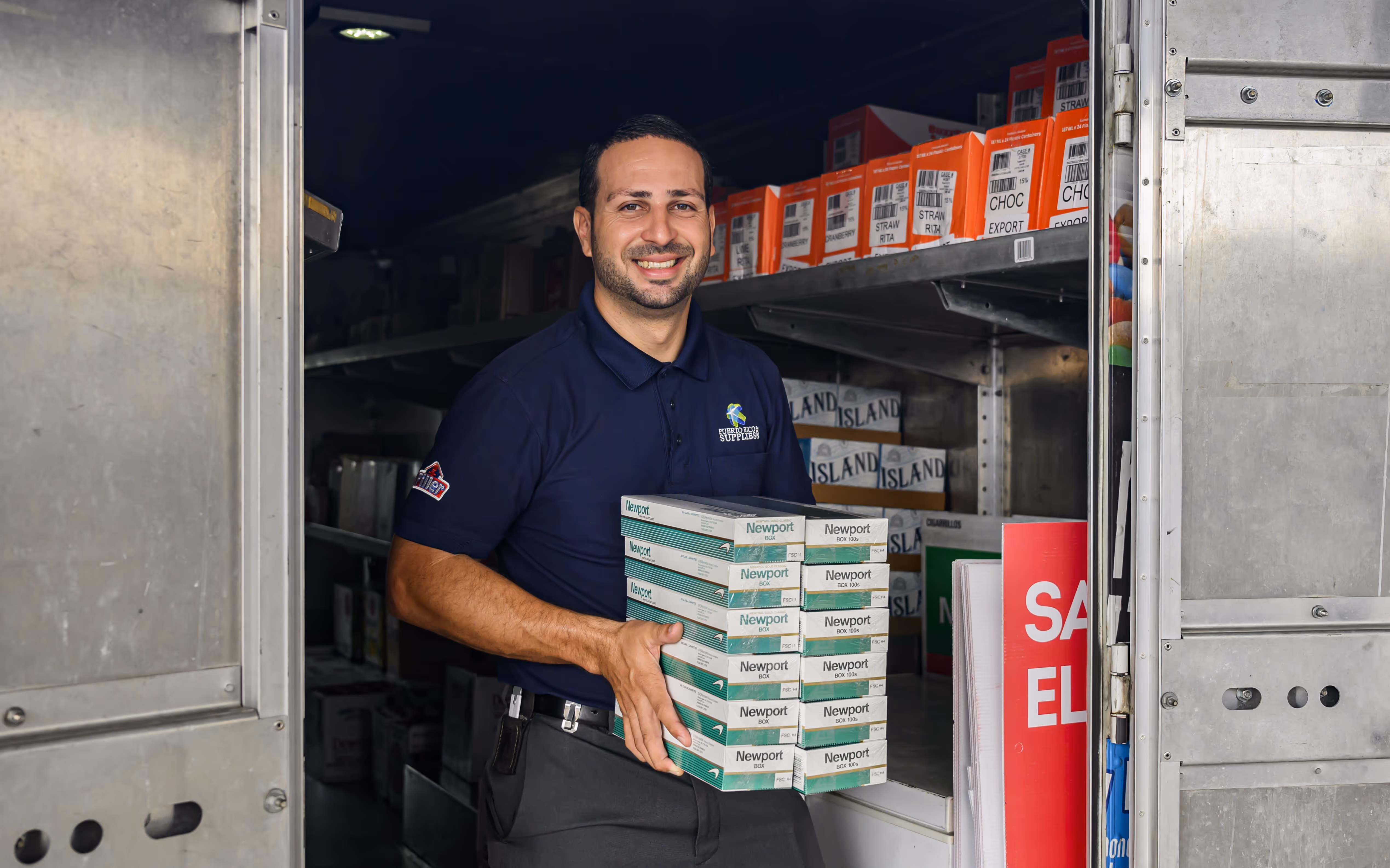 Smiling man in navy blue shirt holding stacked boxes of Newport cigarettes inside a metal storage truck.