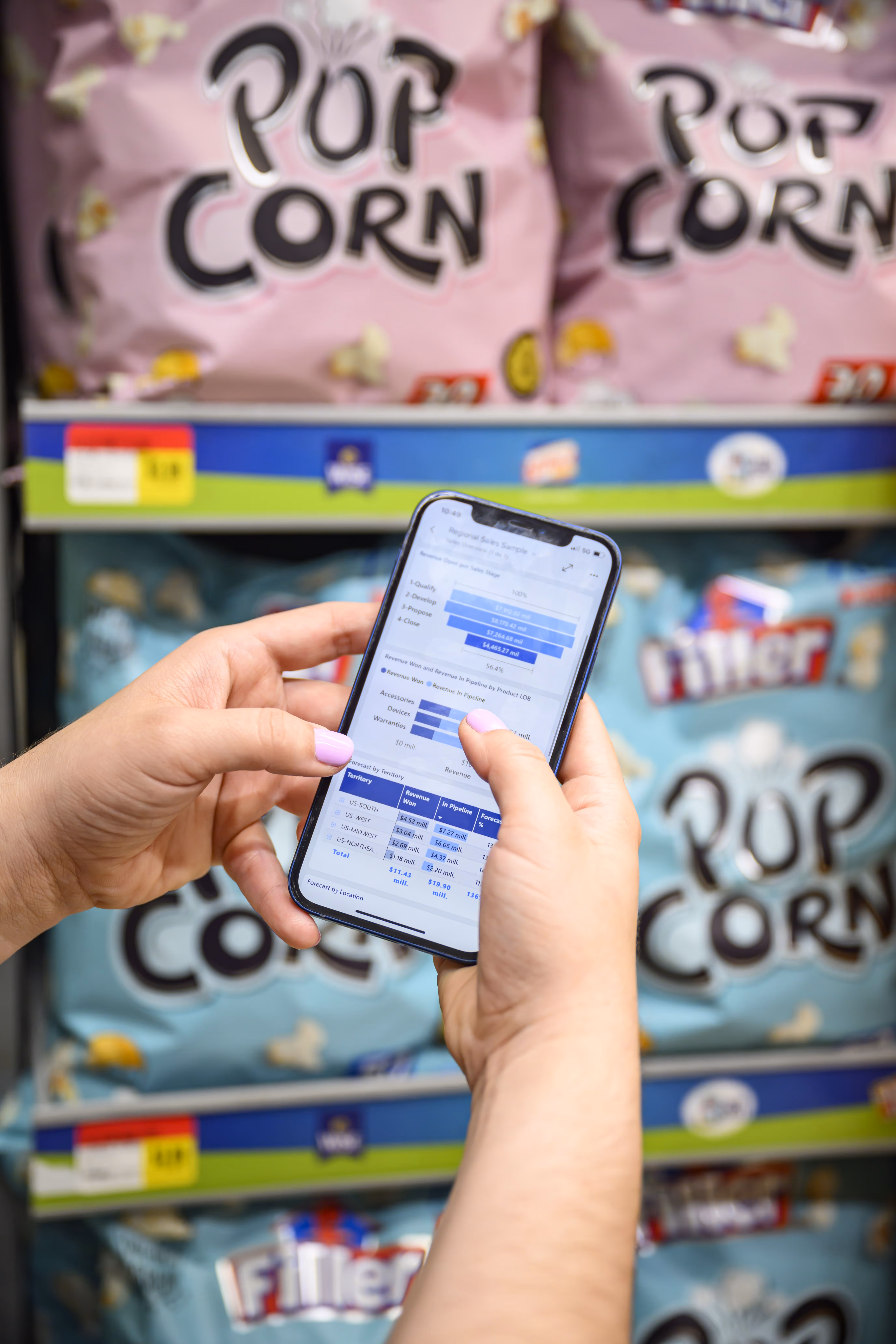 Person holding smartphone showing sales data in front of supermarket shelves stocked with popcorn bags.