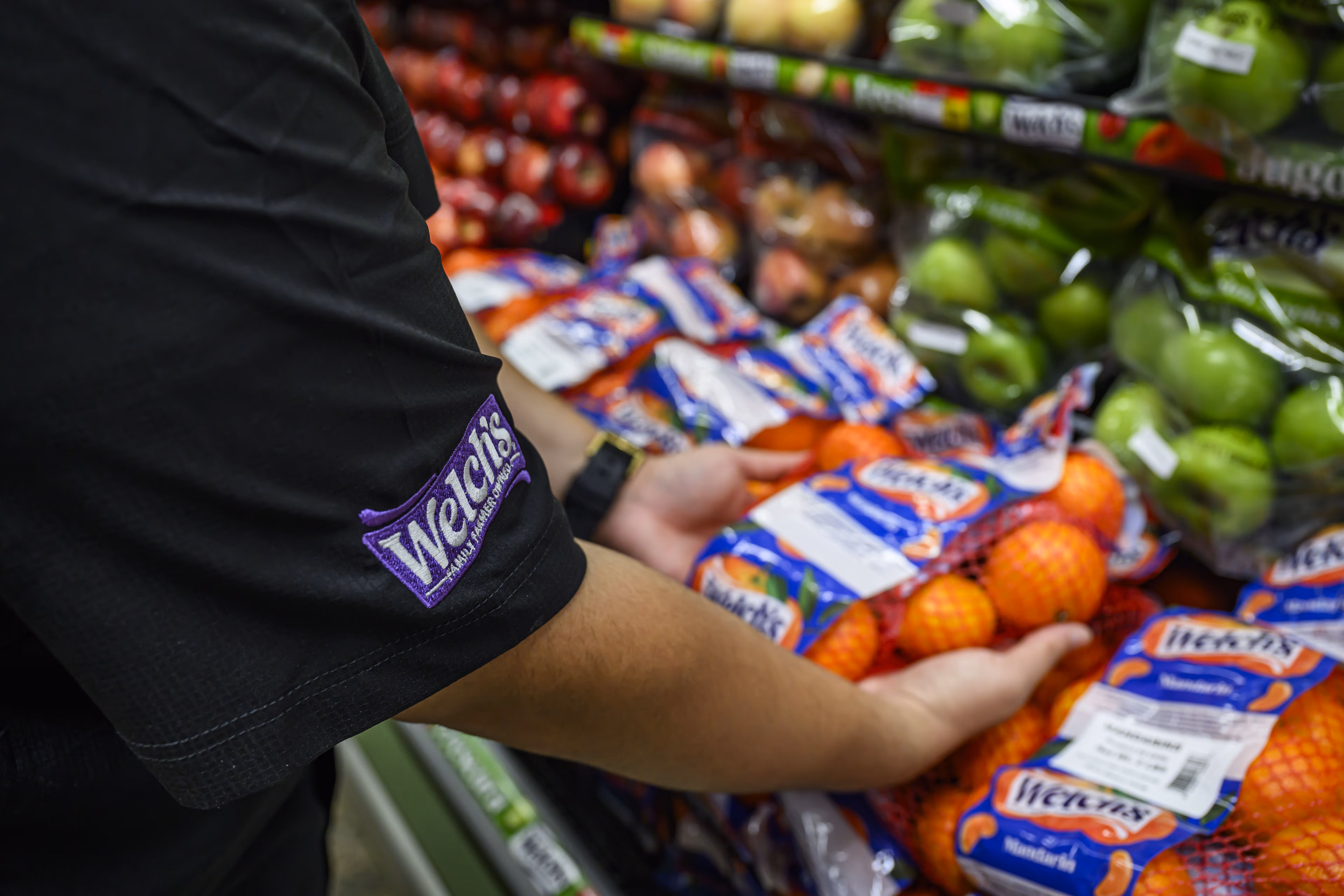 Person wearing a black shirt with a Welch's logo on the sleeve arranging bags of Welch's mandarins in a grocery store produce section.