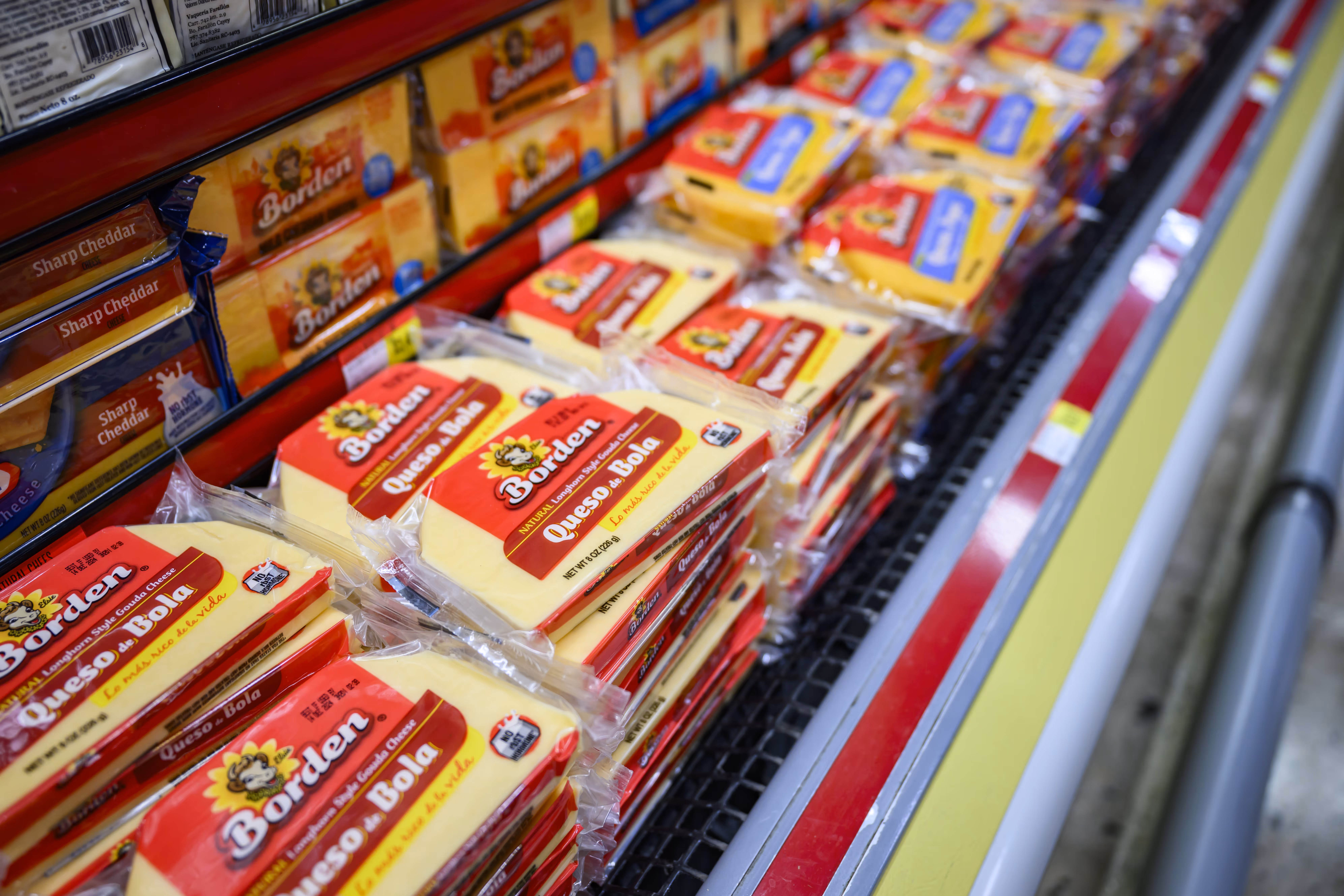 Packages of Borden Queso Bola natural longhorn style Gouda cheese stacked in a supermarket refrigerated display next to sharp cheddar cheese.