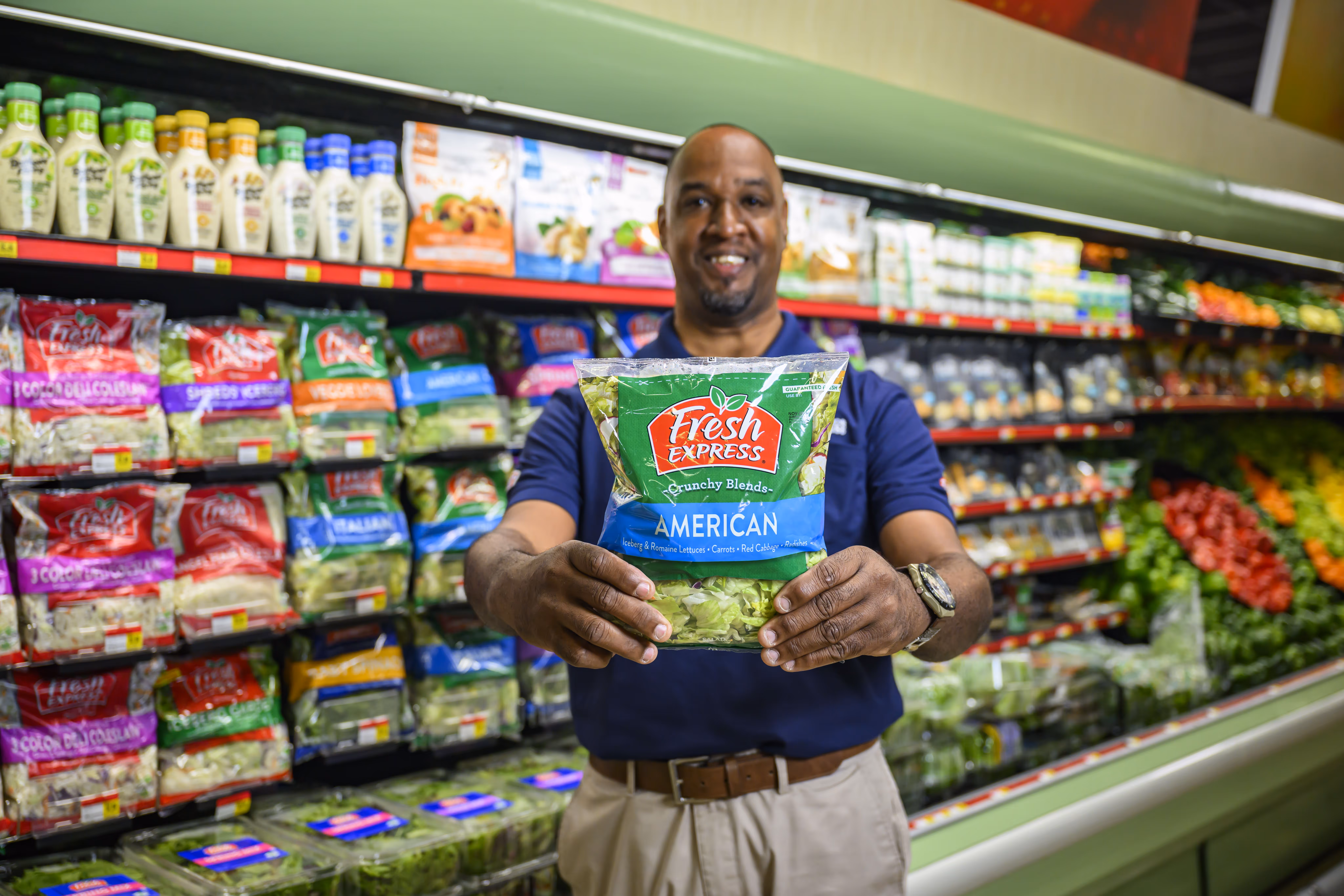Man in a navy blue shirt holding a bag of Fresh Express American salad mix in a grocery store produce aisle.