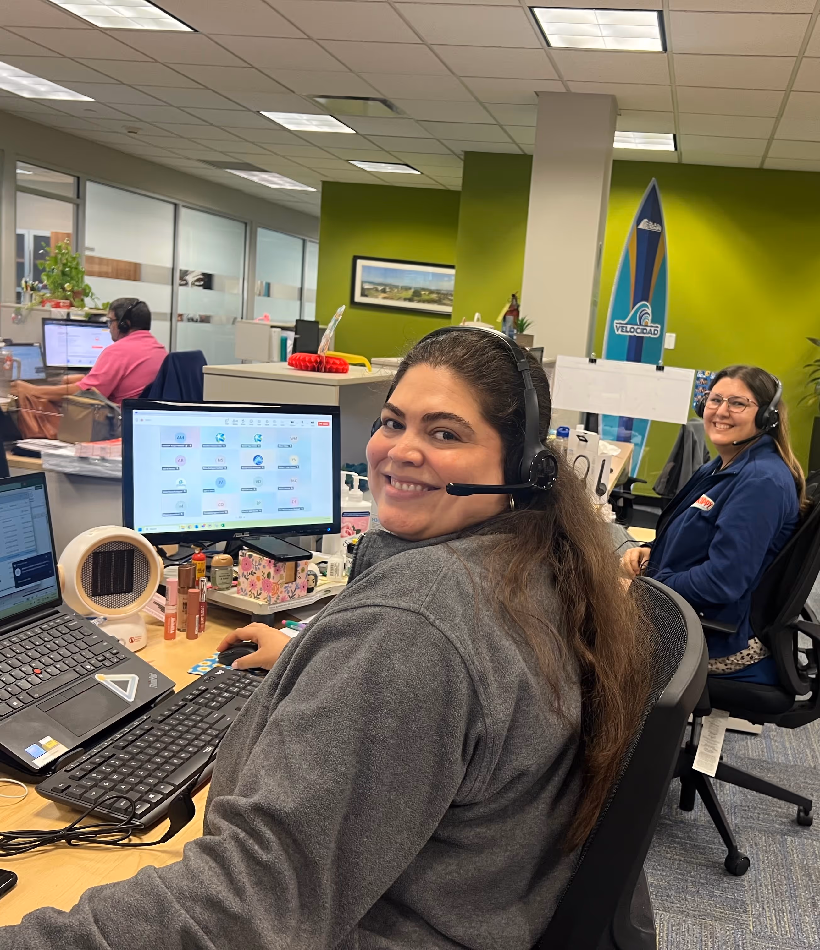 Two smiling women wearing headsets working at desks with computer monitors in a modern office with green walls.