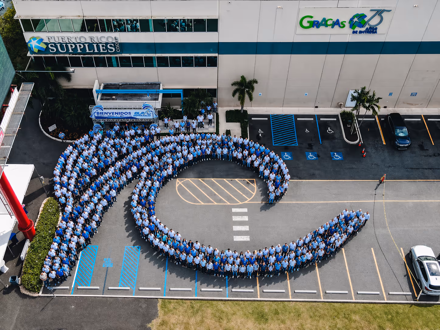 Large group of people wearing blue shirts forming a human wave shape in a parking lot outside a building with Puerto Rico Supplies Corp and Gracias 75 Años de Entrega signs.