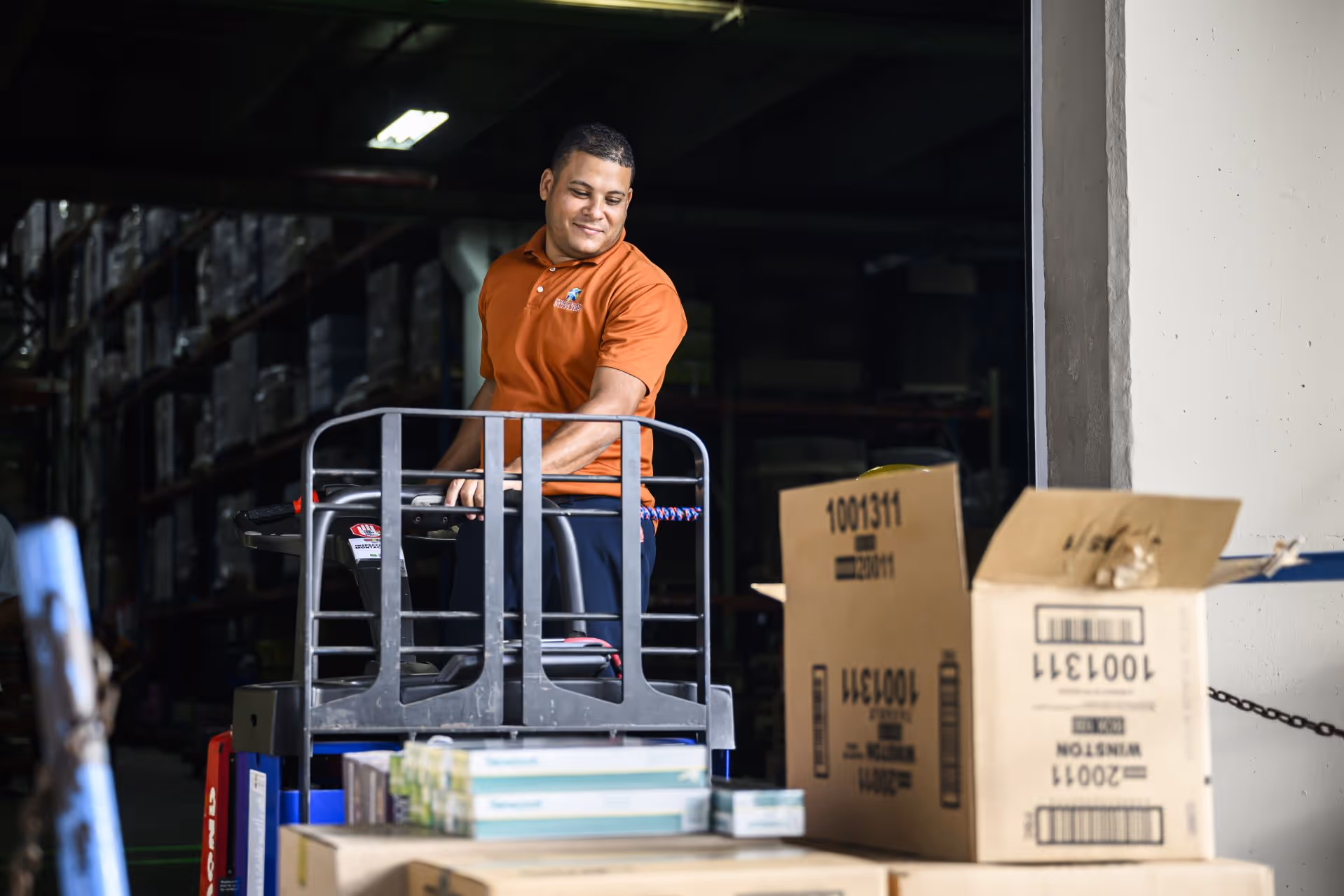 Warehouse worker in an orange shirt operating a pallet jack near stacked cardboard boxes.