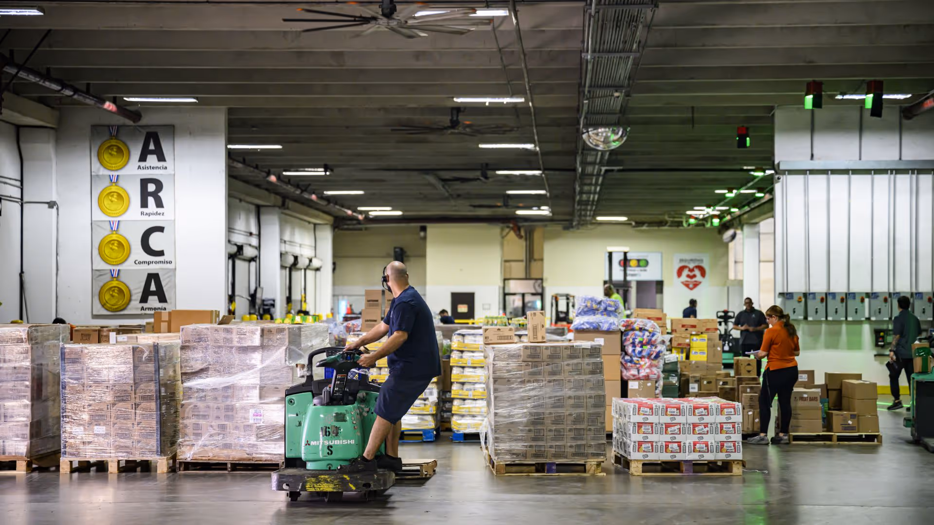 Warehouse worker operating a pallet jack among stacks of boxed and wrapped goods on pallets.