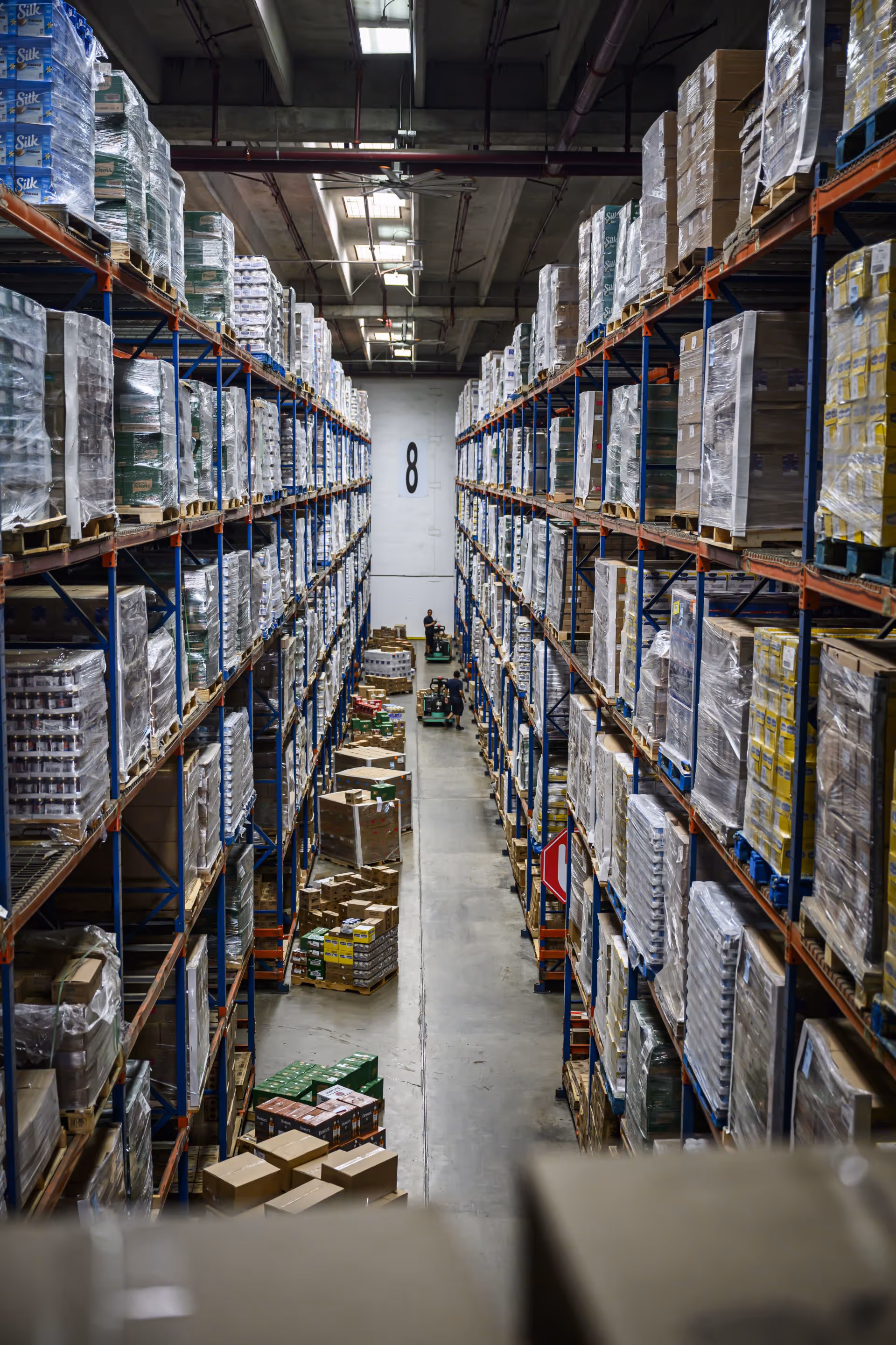 Warehouse aisle with tall shelves stocked with palletized goods and two workers on forklifts in the distance.