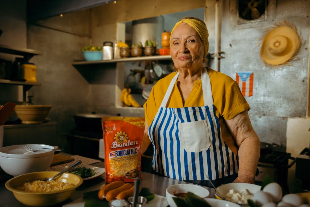 Elderly woman in yellow and striped apron standing in a kitchen with Puerto Rican flag on the wall.