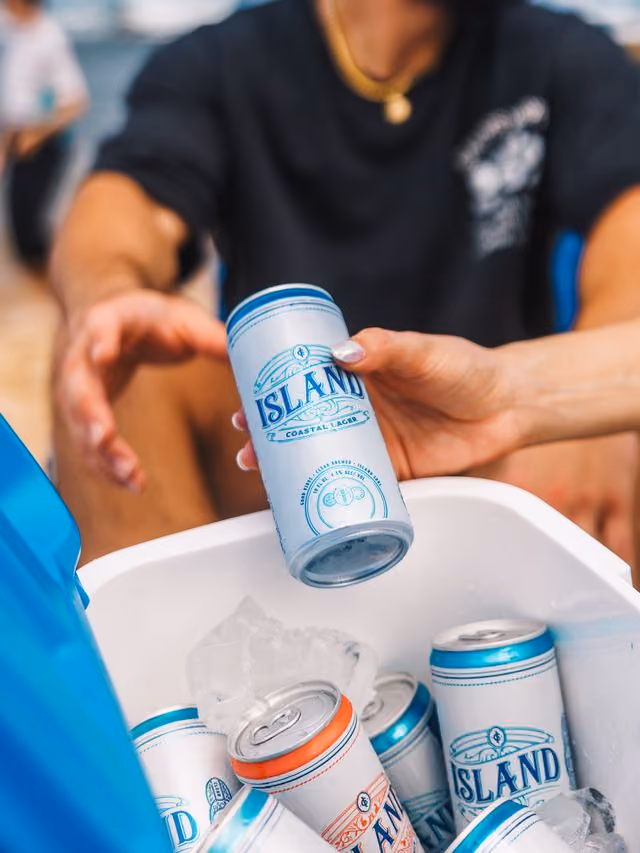 Hand holding a can of Island Coastal Lager beer over an ice-filled cooler with other cans inside.