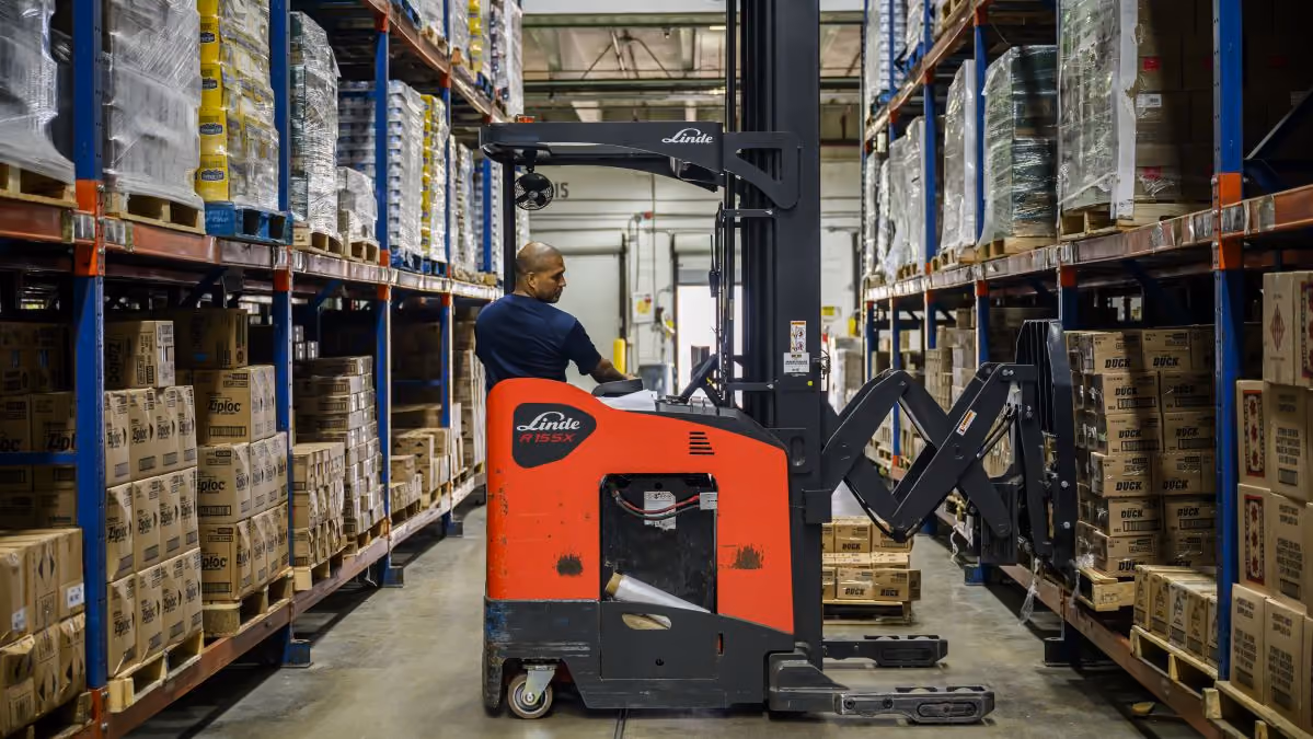 Warehouse worker operating a red forklift, moving boxes on shelves in an aisle.