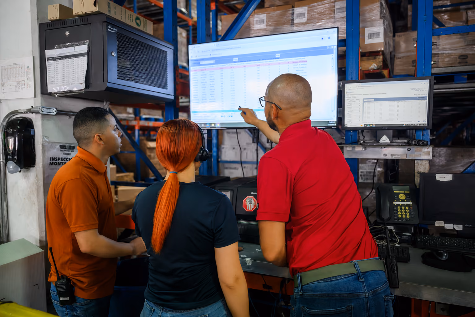 Three warehouse workers study data on two large monitors mounted on blue shelves.