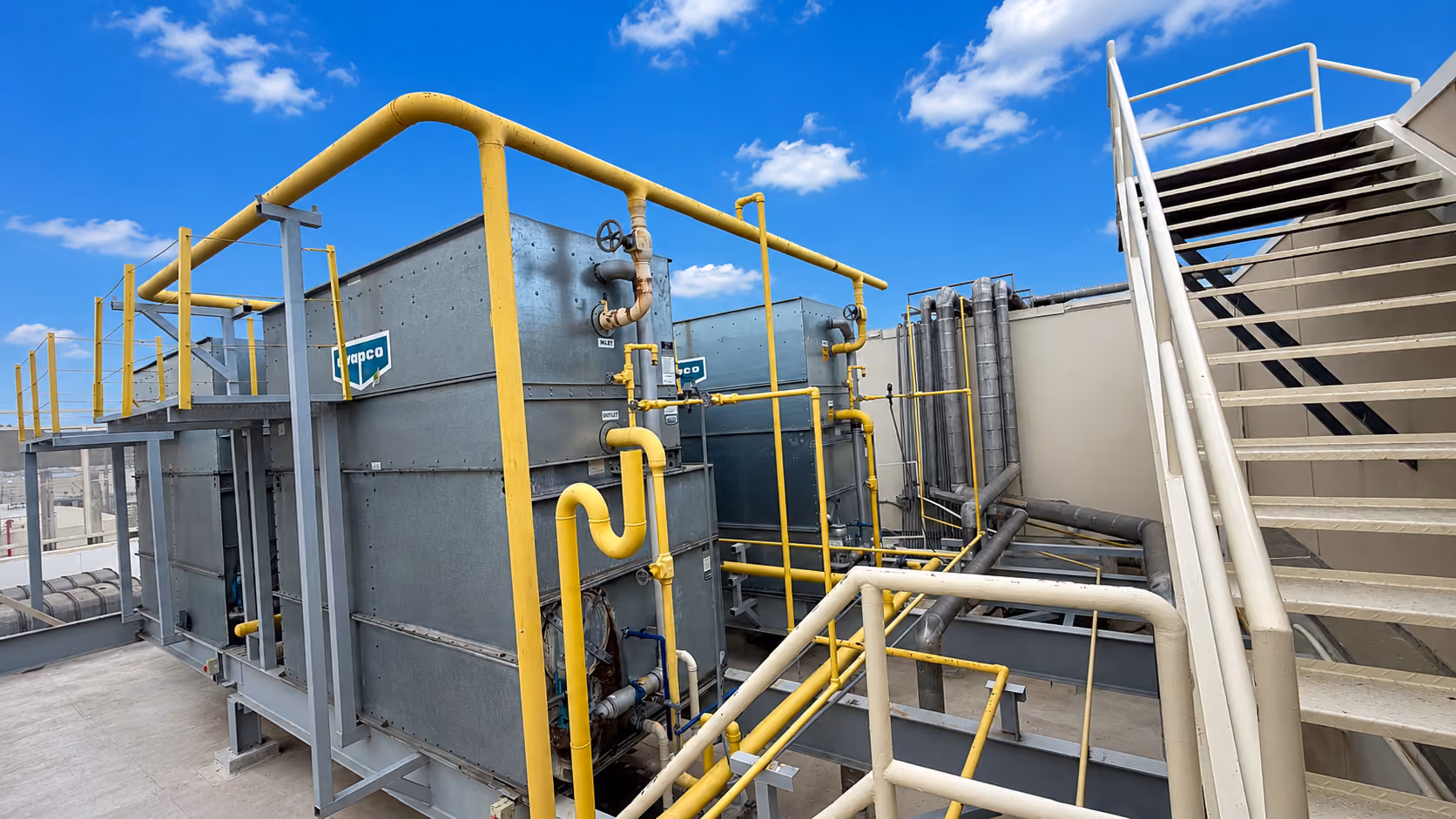 Industrial rooftop with large metal machinery, yellow piping, and metal stairway against a blue sky.