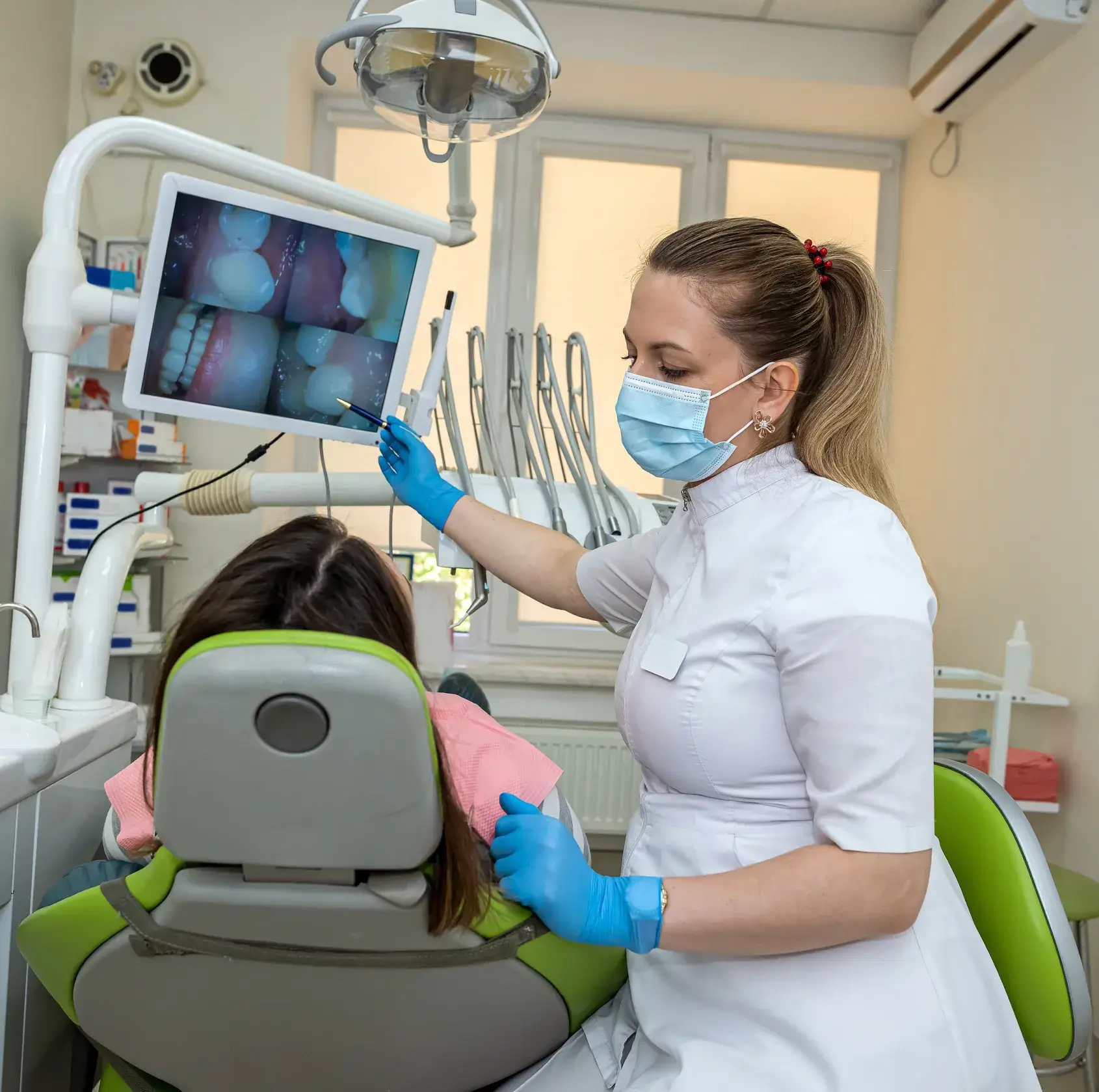 Dentist wearing a mask and gloves pointing at dental X-rays on a monitor while examining a seated patient in a dental clinic.