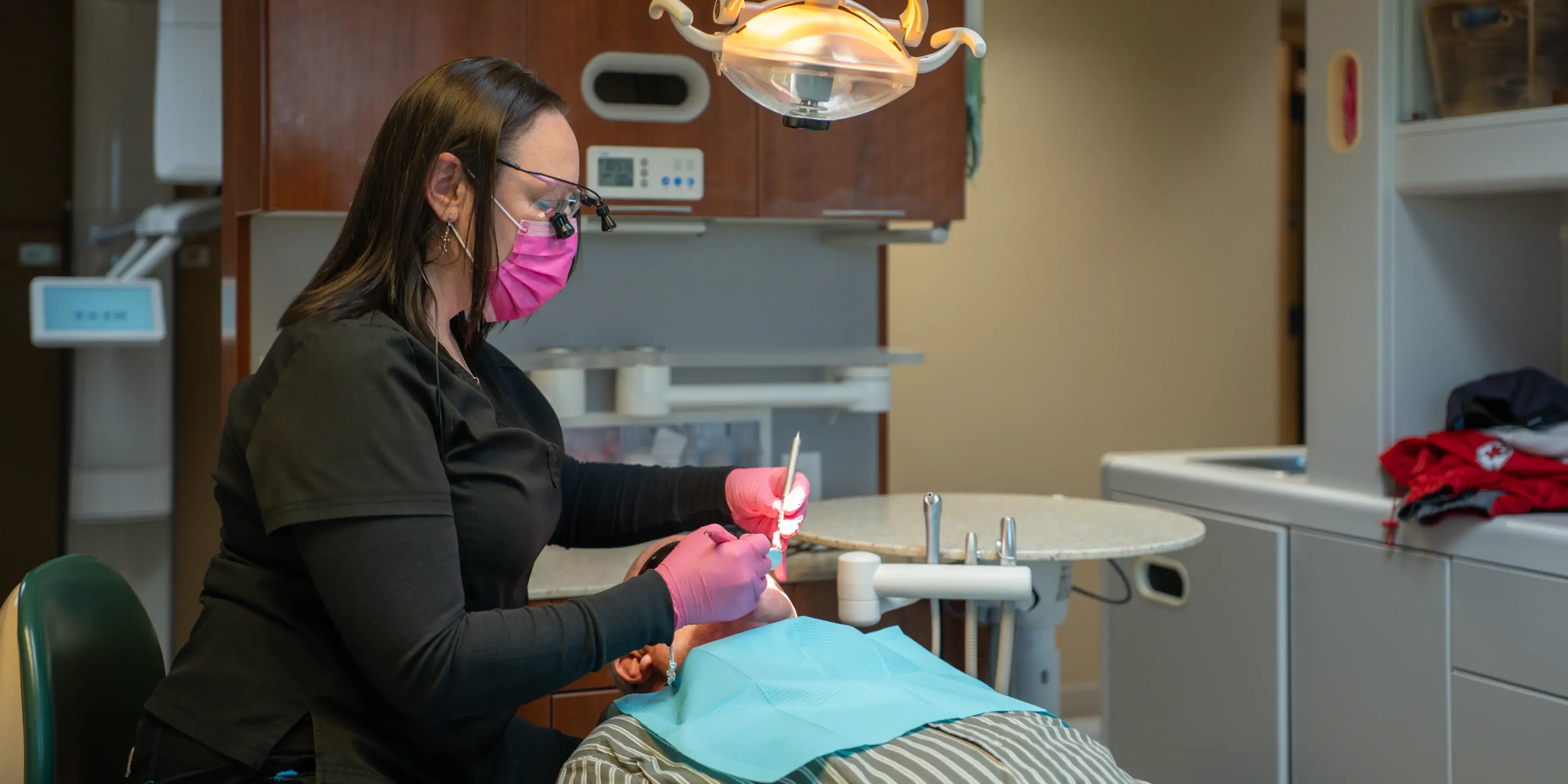 Dental professional wearing a pink mask and gloves treating a patient lying back in a dental chair.