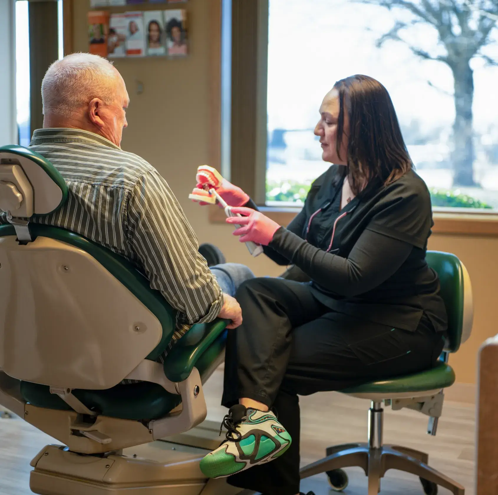 Dental professional demonstrating teeth cleaning using a model to an older male patient seated in a dental chair.