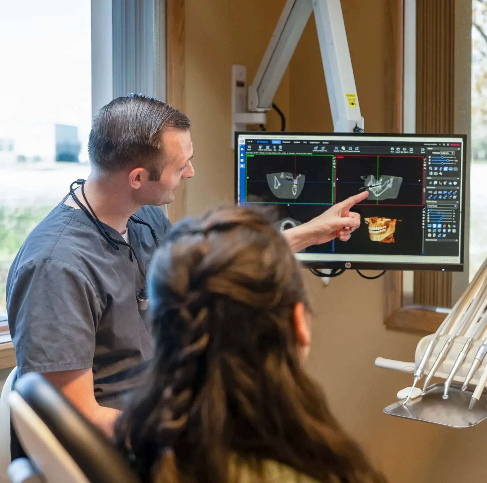 Dentist pointing to dental X-ray images of teeth on a computer screen while explaining to a patient in a clinic.