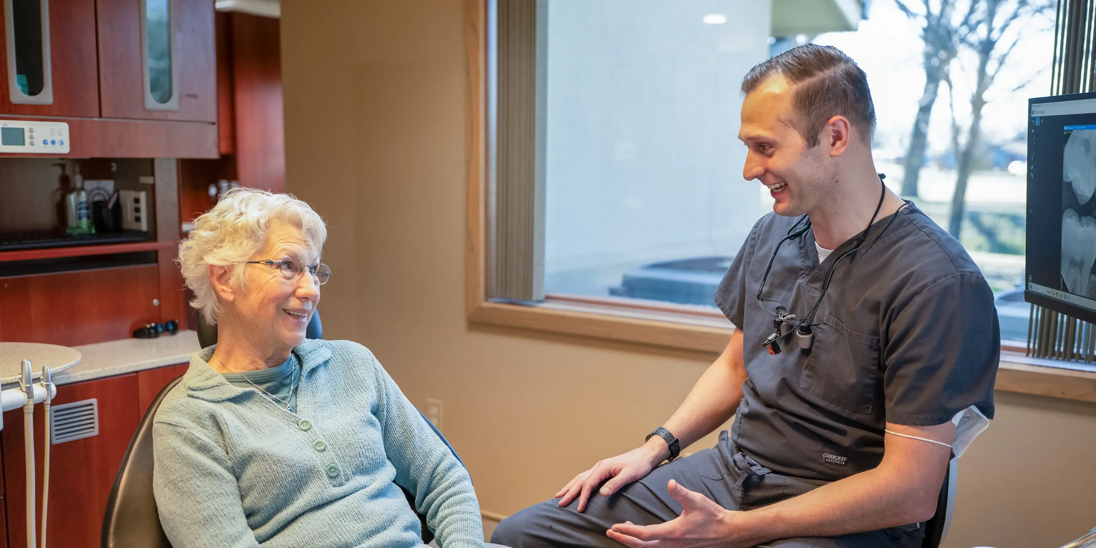 Smiling dentist in scrubs talking with a seated elderly female patient in a dental office.