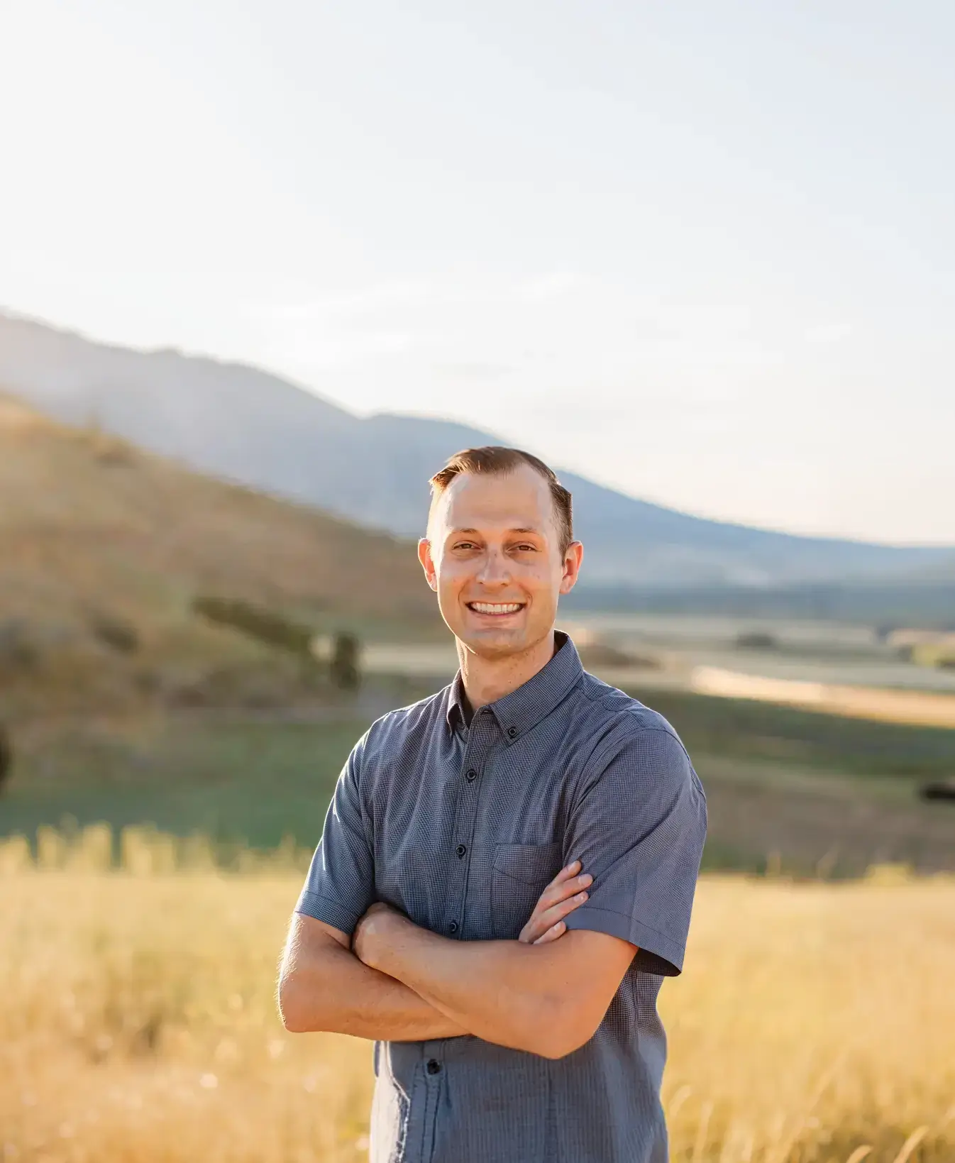 Smiling man in a blue short-sleeve shirt with arms crossed standing in a sunlit field with mountains in the background.