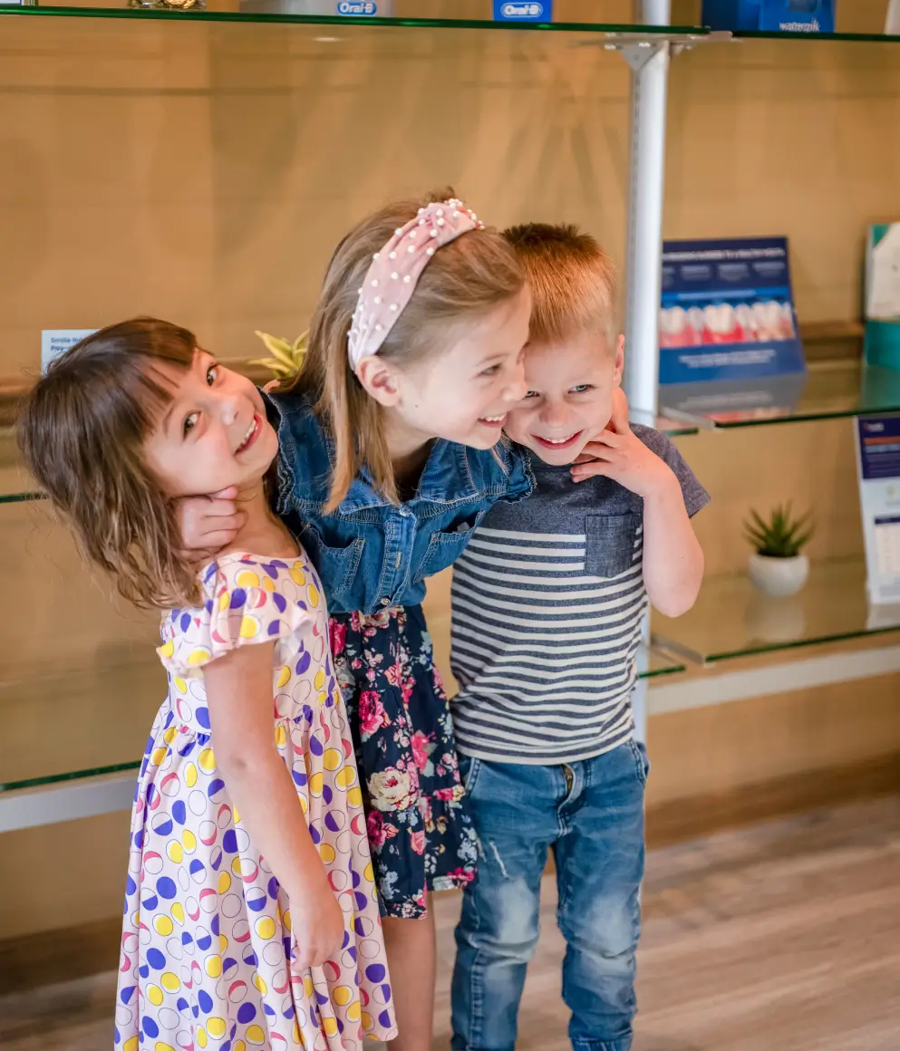 Three young children smiling and hugging each other in a room with glass shelves and dental care products.