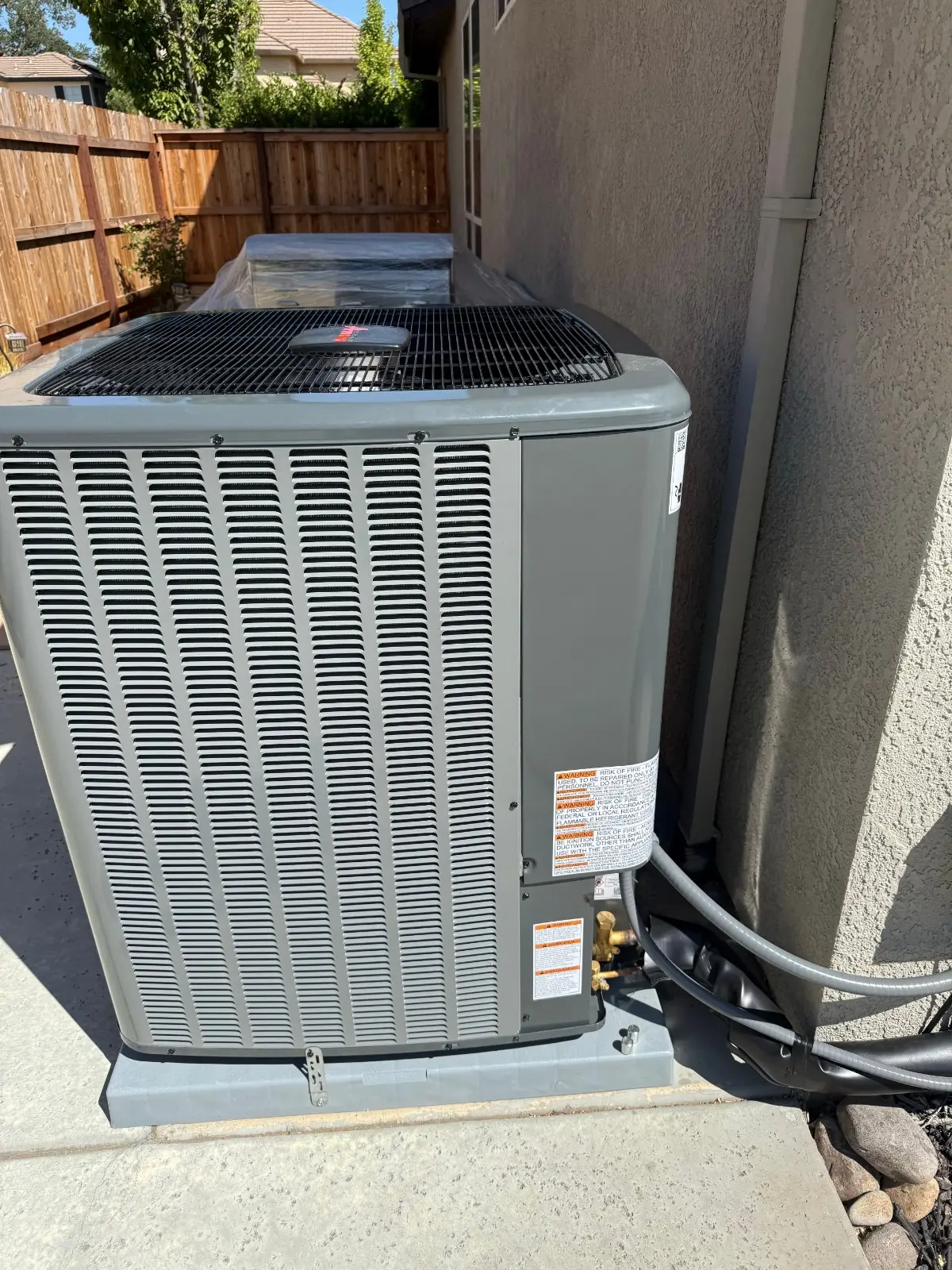  A large, dark grey outdoor air conditioning condenser unit stands on a grey plastic pad next to a light-colored stucco wall. It features slatted sides and a fan visible through the top grille. Refrigeration lines and electrical conduits are connected near the base. A wooden fence is visible in the background.