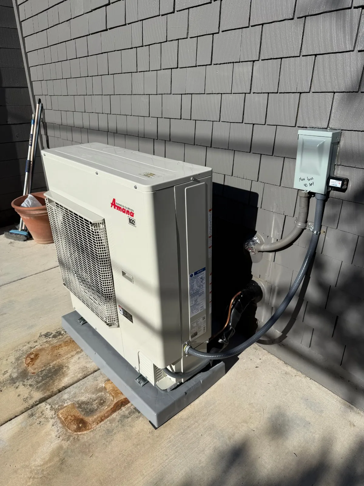 A new, light gray, vertical Amana brand heat pump or AC condenser unit is installed on a dark gray pad against a residential wall covered in dark gray shingle siding. It is connected by conduits to an electrical disconnect box mounted on the wall, which has handwritten text indicating 'Max fuse 40 HP'.