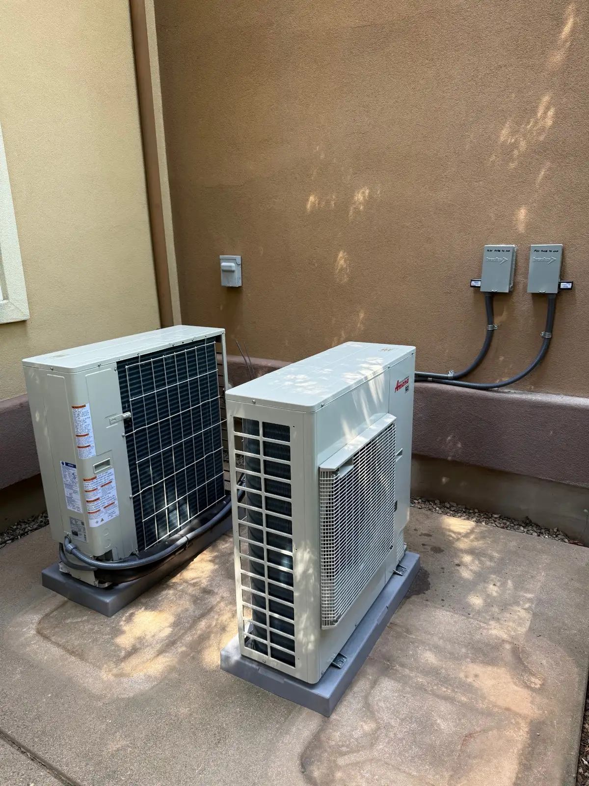 Two rectangular, off-white mini-split heat pump or air conditioning condenser units are installed on grey plastic pads on a concrete patio. They sit against a plain brown stucco wall. The unit in the foreground is oriented vertically, while the unit behind it is horizontal. Two electrical disconnect boxes are mounted high on the wall, connected to the units by black conduits.
