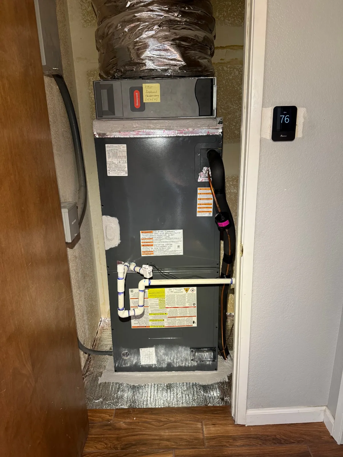  A dark gray, rectangular air handler unit is installed vertically in a small closet with a wood-grain floor. A large, insulated flexible duct is connected to the top of the unit. White PVC condensate lines with a safety switch are connected near the bottom. To the right, a white wall holds a modern, black digital thermostat displaying "76."