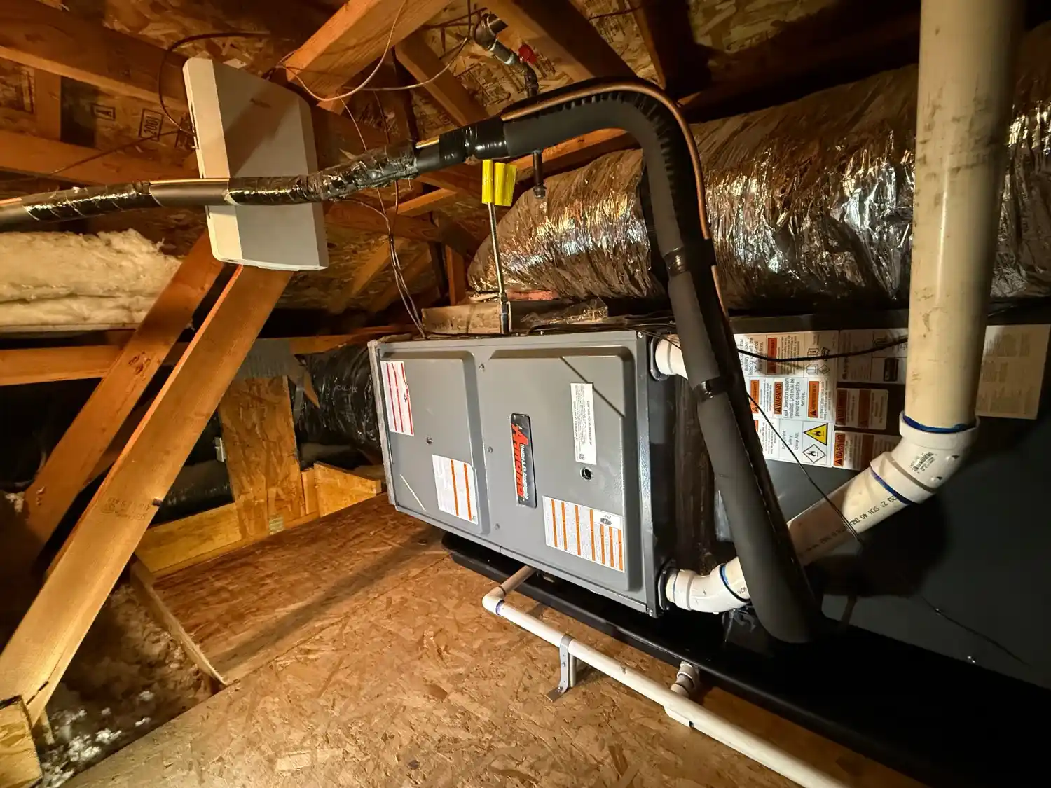 A gray horizontal air handler unit is installed in an unfinished attic space, resting on plywood flooring. It features a visible Amana logo. Insulated black refrigerant lines curve over the unit, connecting to the coil section. Large, white PVC piping for condensate drainage and a flexible metallic air duct are also visible, surrounded by wooden framing and fiberglass insulation.
