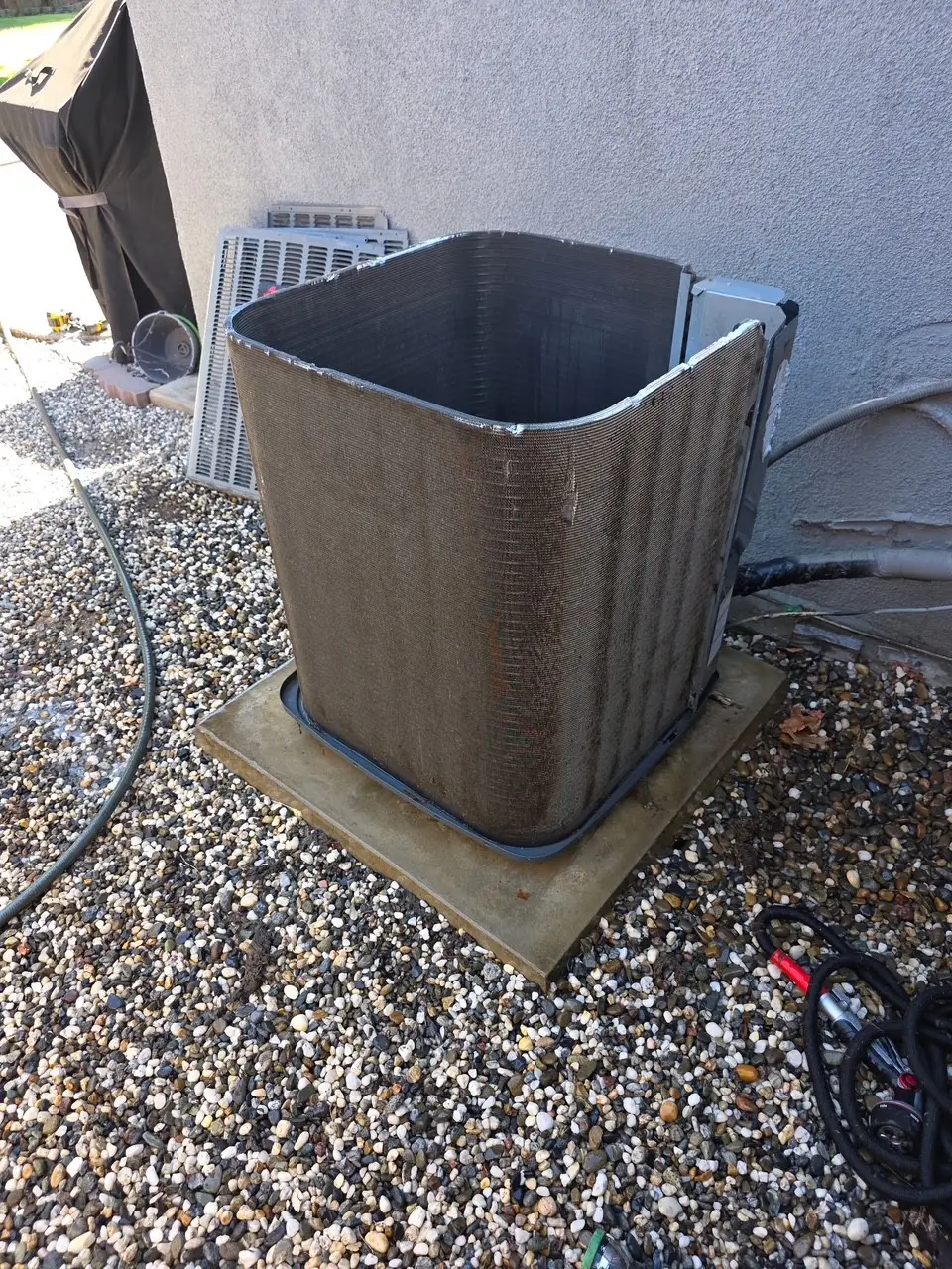 condenser coil of an outdoor air conditioning unit (or heat pump) with the protective metal cage removed, exposing the dirty coil fins. The unit is resting on a concrete pad on a bed of gravel, next to a building wall. Tools and the removed casing are visible nearby, indicating a maintenance or cleaning task.