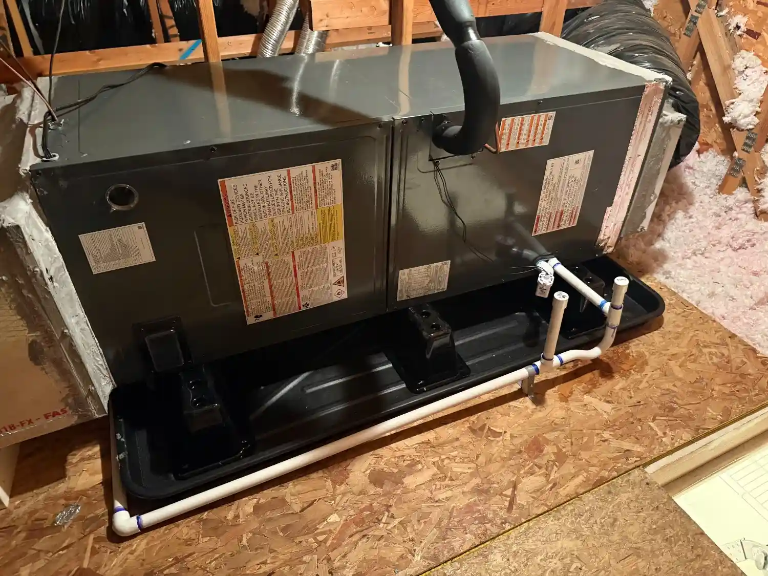 An HVAC air handler unit in an attic, sitting in a black drain pan with white PVC condensate lines attached.