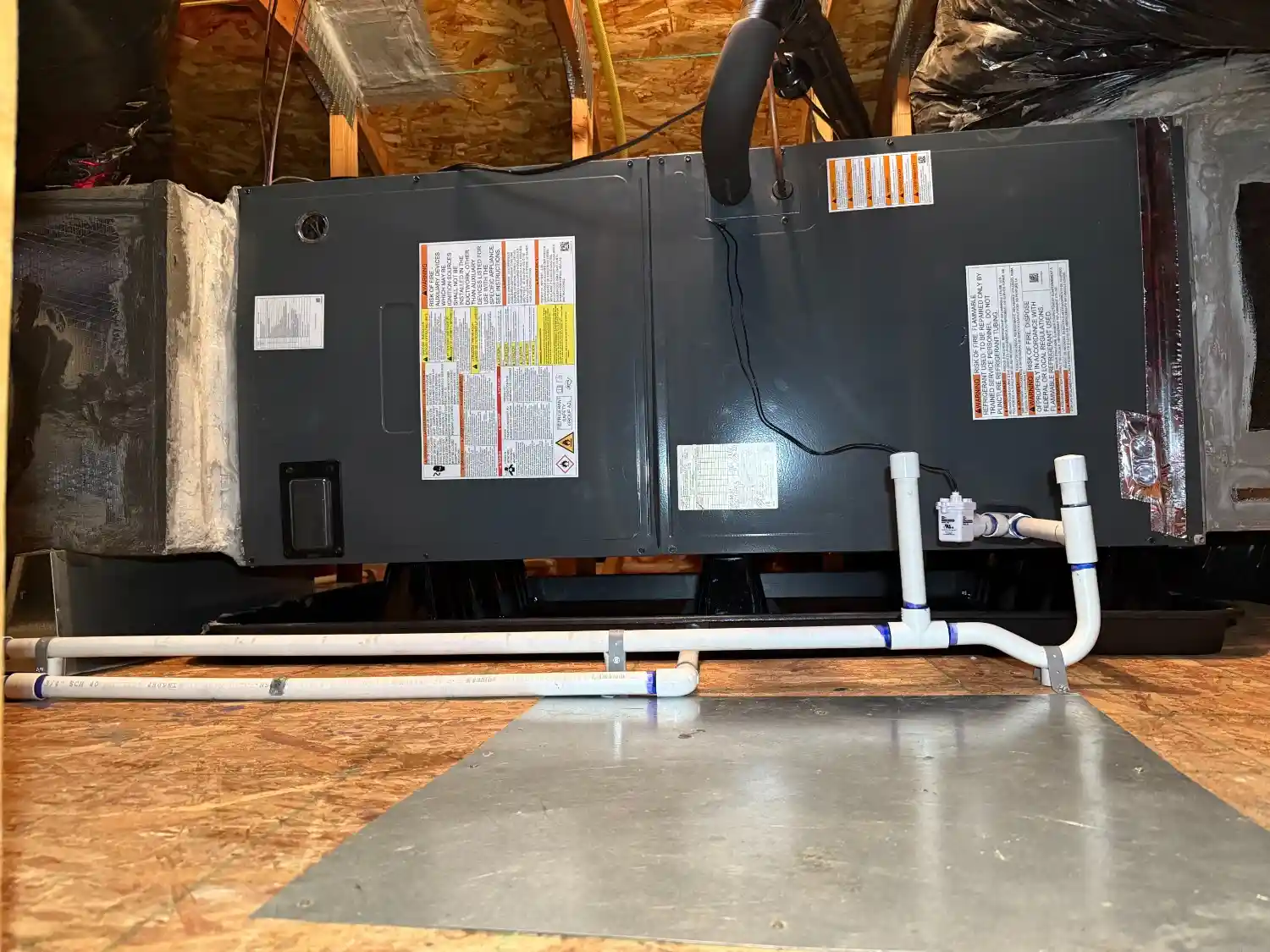 An HVAC air handler unit in an attic, showing the unit, PVC condensate drain pipes, and a metal drip pan extension.