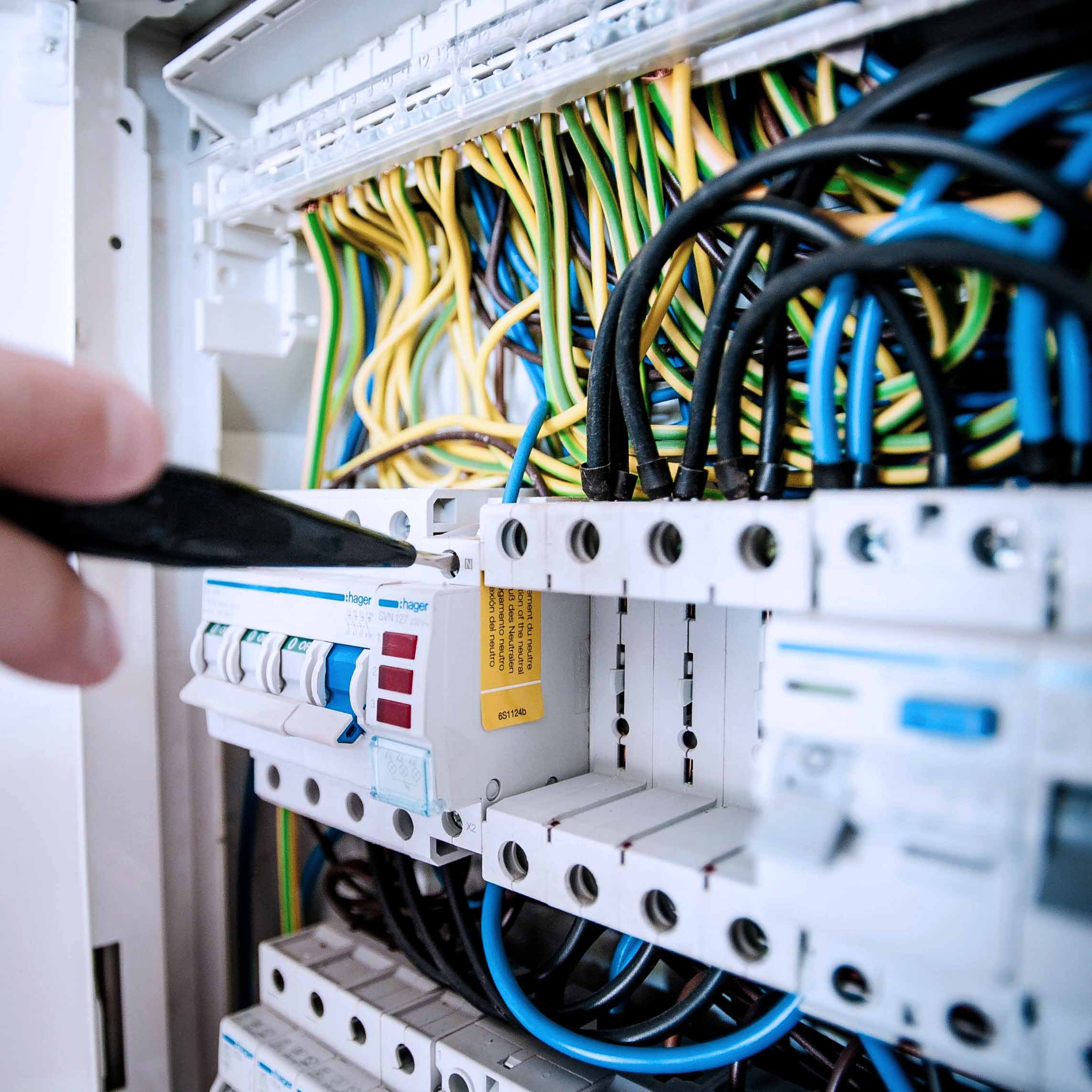 Hand hält Schraubenzieher und arbeitet an einem elektrischen Sicherungskasten mit farbigen Drähten.