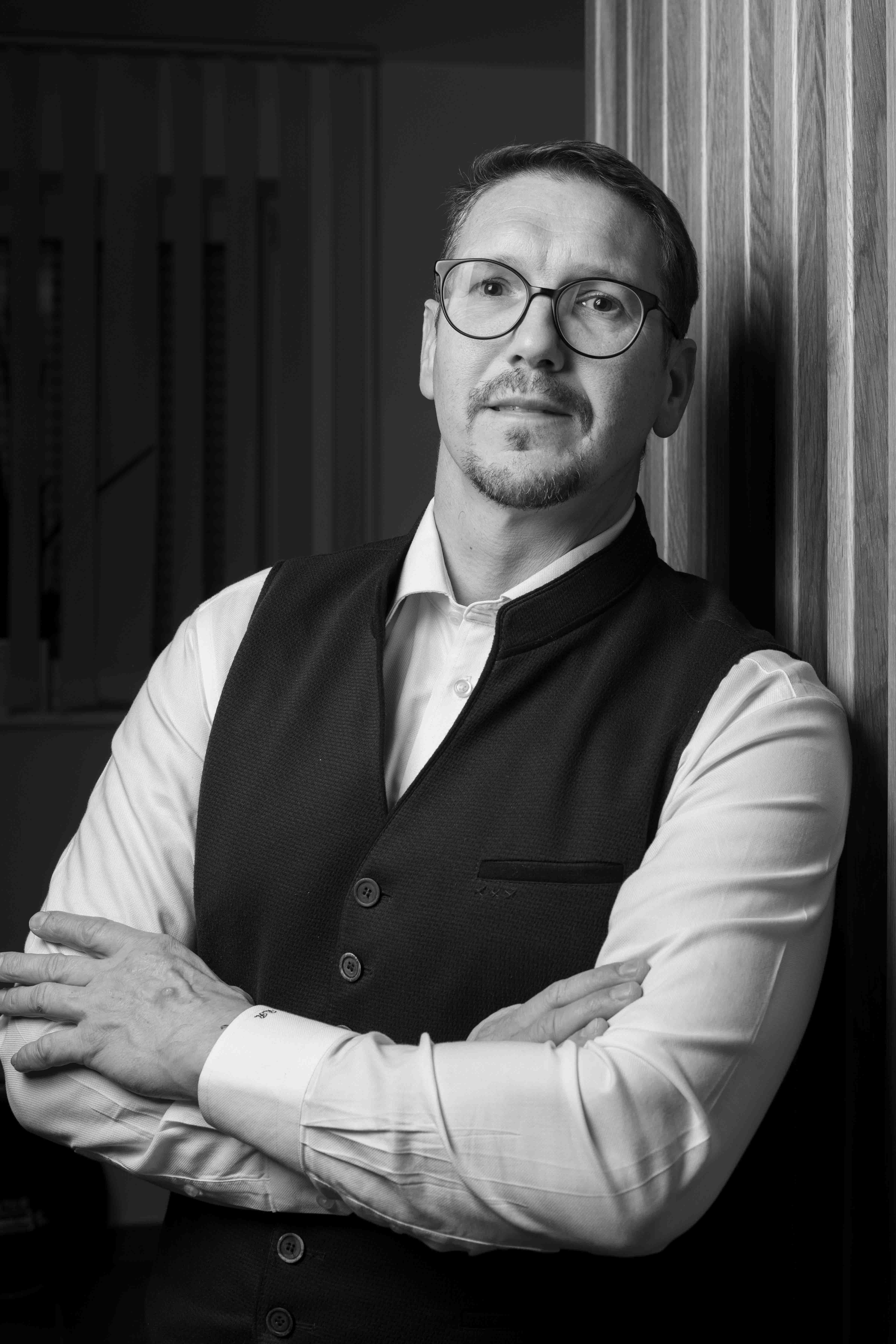 Schwarz-weiß-Porträt eines Mannes mit Brille, der mit verschränkten Armen an eine Holzwand lehnt.