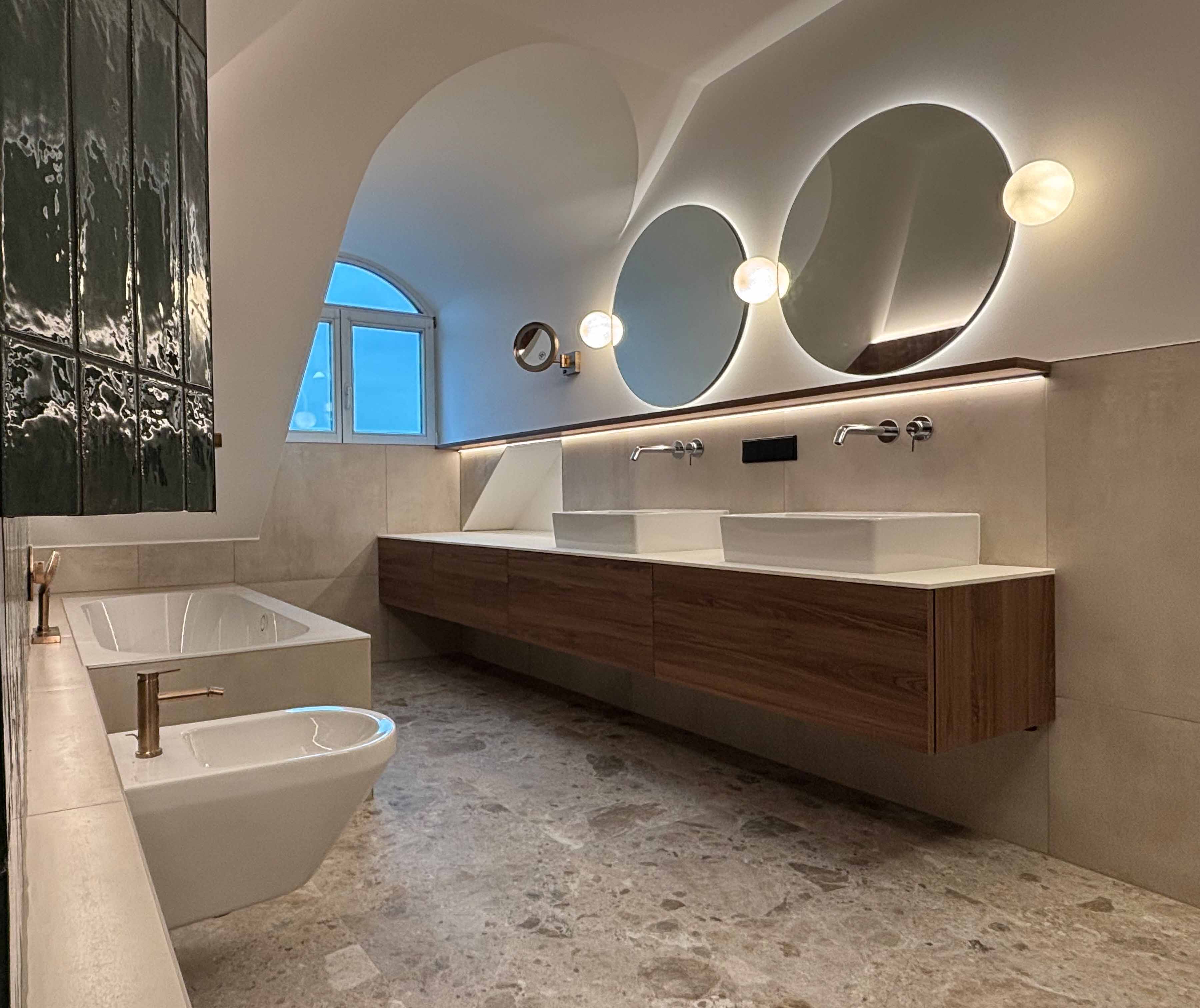 Modernes Badezimmer mit zwei runden beleuchteten Spiegeln, zwei Waschbecken, einer Badewanne und einem Bidet vor einer grauen Fliesenwand und Marmorboden.