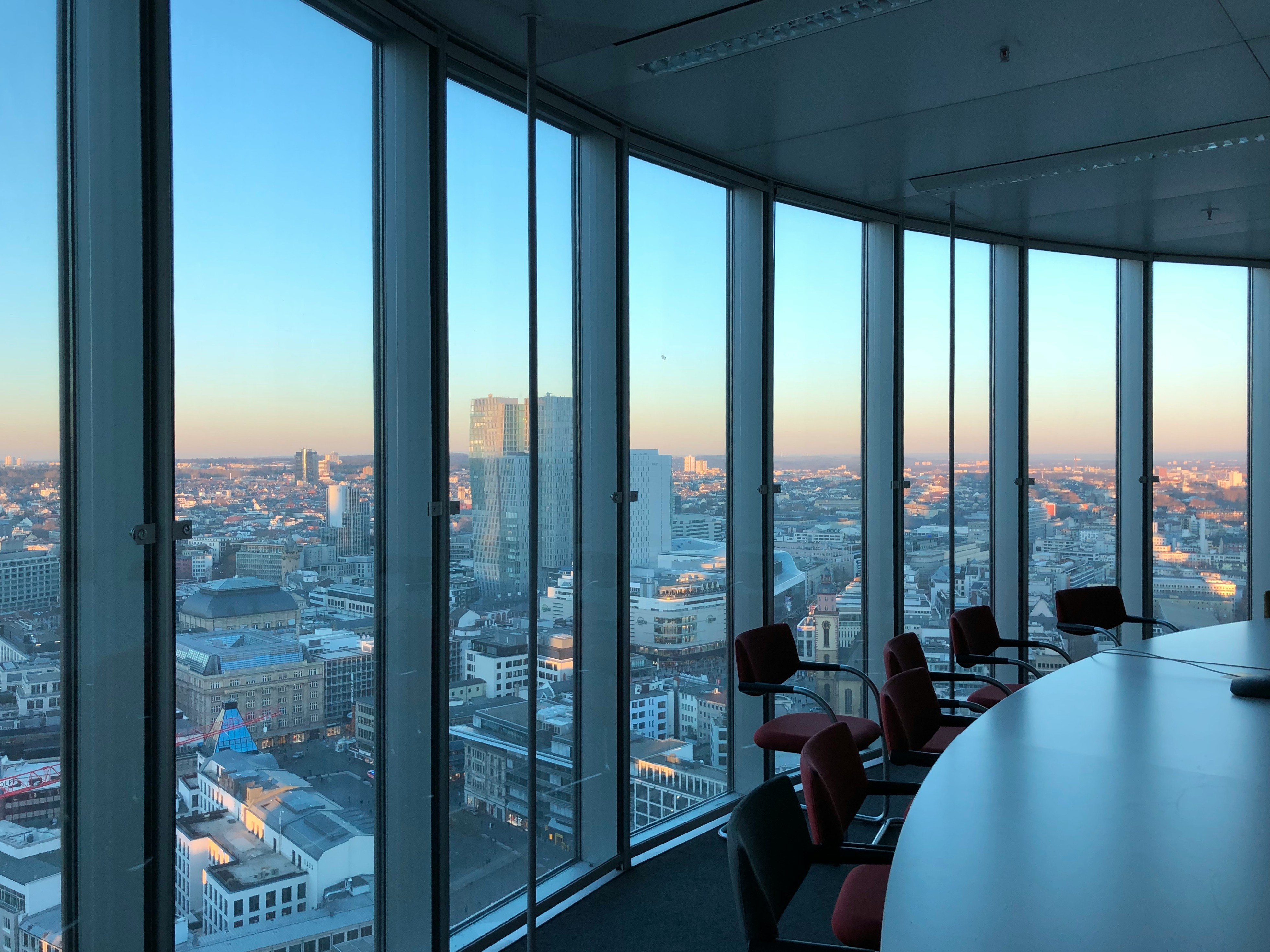 City skyline view through tall office windows overlooking buildings during sunset with empty conference table and red chairs.