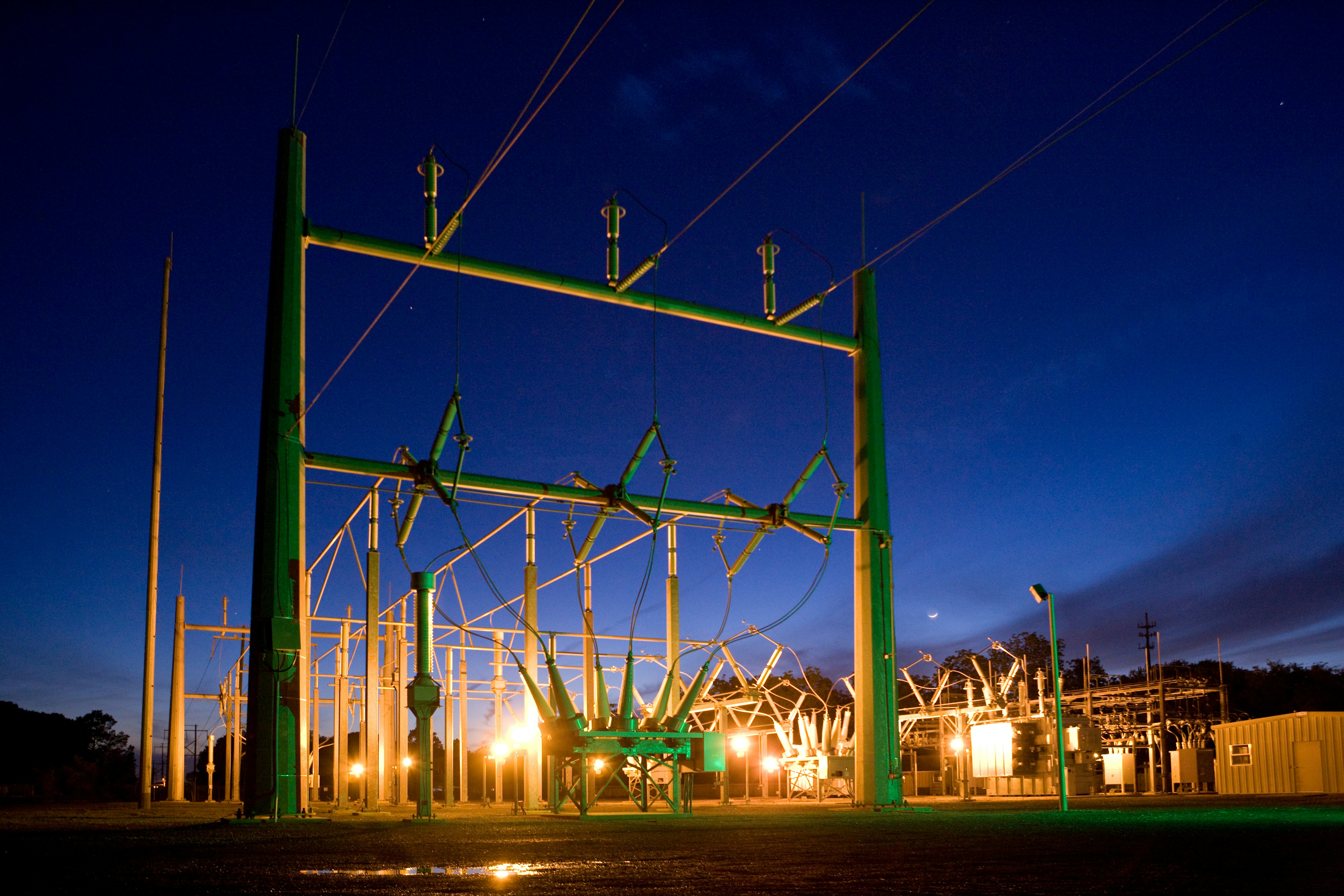 Electrical substation with illuminated equipment and power lines at dusk under a dark blue sky.