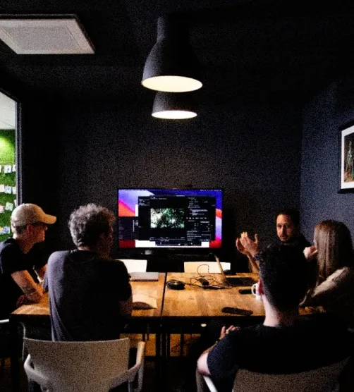 Groupe de cinq personnes assises autour d'une table en bois dans une salle sombre, regardant un grand écran d'ordinateur.