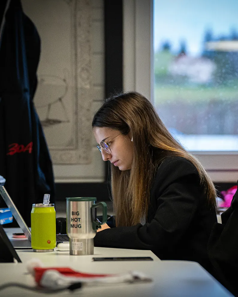 Jeune femme portant des lunettes travaillant sur un ordinateur portable à une table avec deux gourdes et une grande tasse métallique.