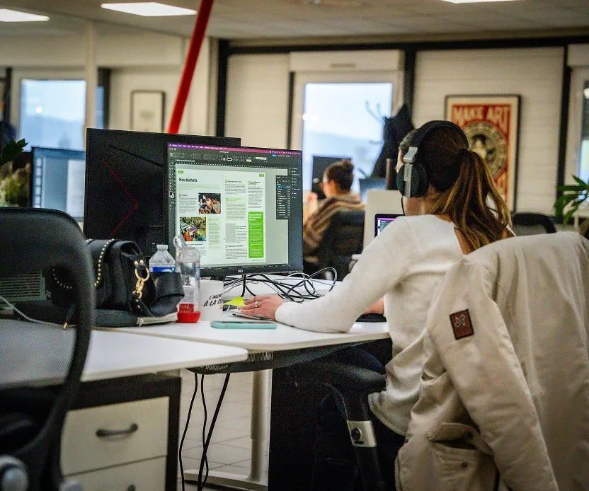 Personne portant un casque et travaillant sur un ordinateur dans un bureau avec plusieurs écrans et dossiers.