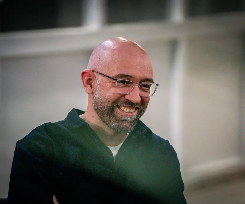 Homme chauve portant des lunettes et une barbe souriant avec un fond flou intérieur.