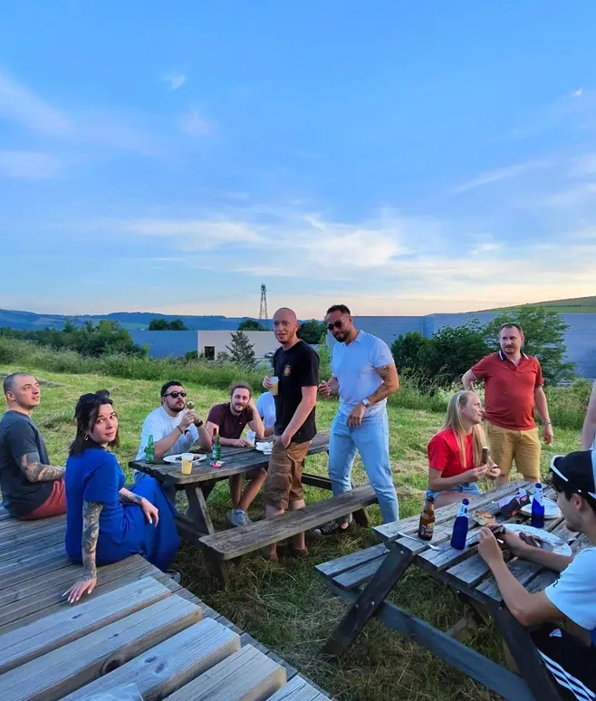 Groupe de personnes assises et debout autour de tables de pique-nique en bois en plein air durant un coucher de soleil.