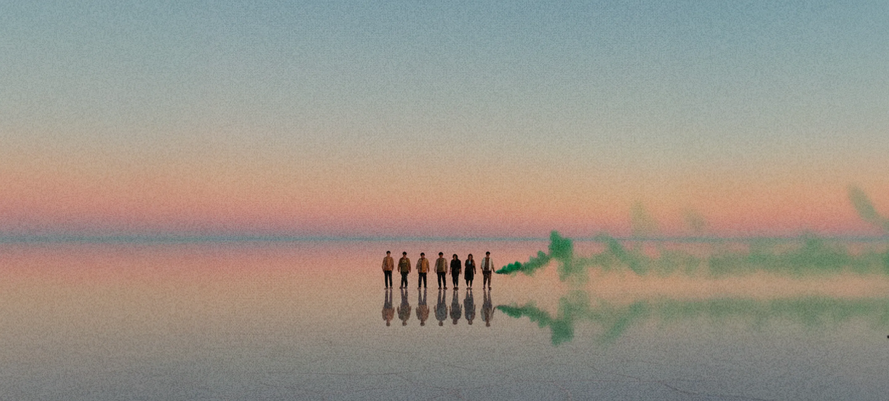 Sept personnes debout côte à côte sur une surface d'eau réfléchissante sous un ciel pastel au coucher du soleil, avec une fumée verte s'échappant d'une d'entre elles.